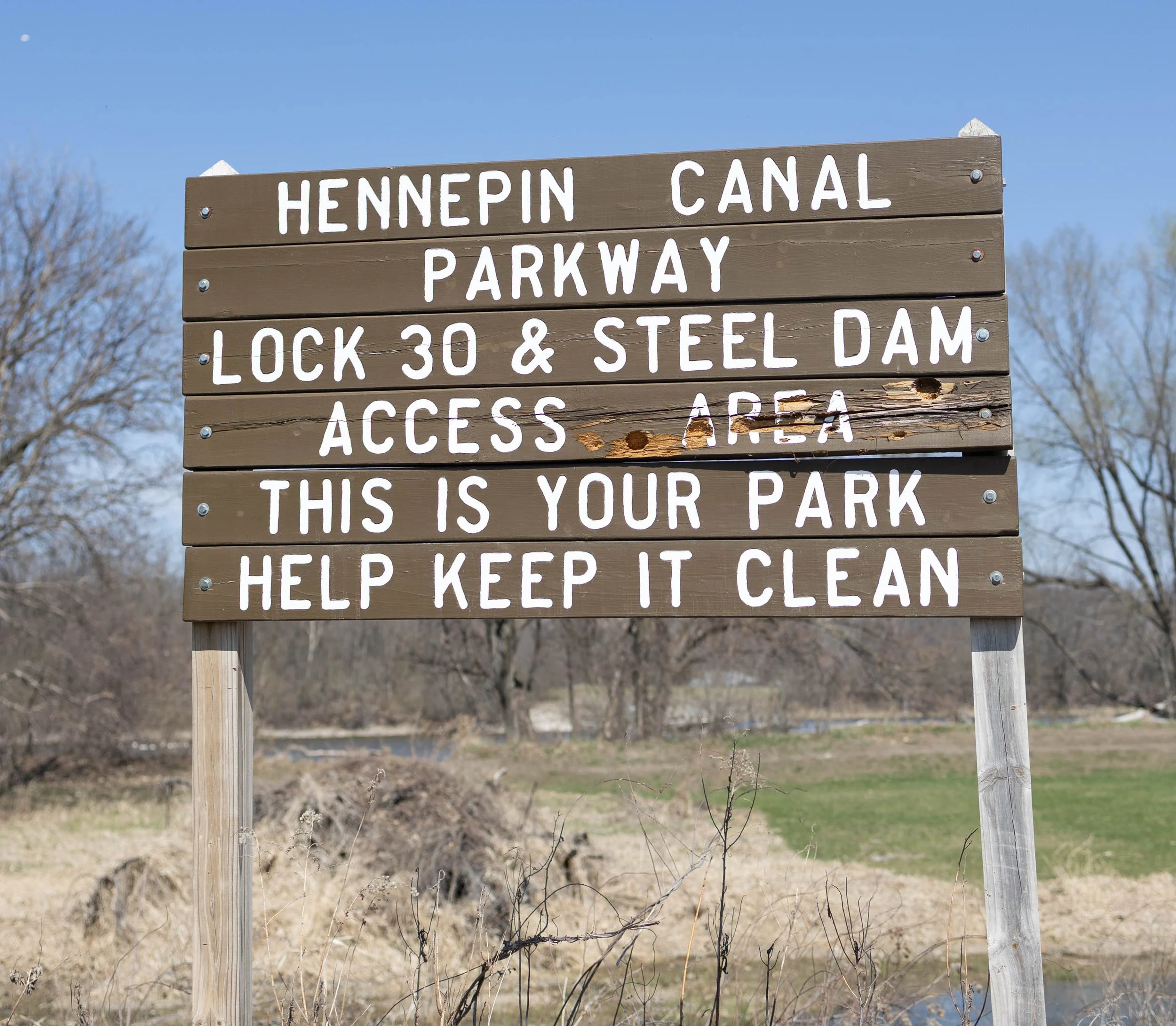 Wooden signboard with white painted text indicating directions and rules at Hennpin Canal Parkway, mentioning lock 30 & steel dam, access area, and reminding visitors to help keep it clean, outdoors with trees and clear sky in background.