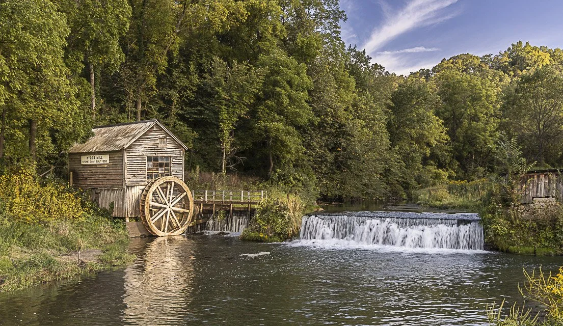A historic watermill with a large wooden wheel beside a small waterfall, surrounded by green trees under a blue sky.
