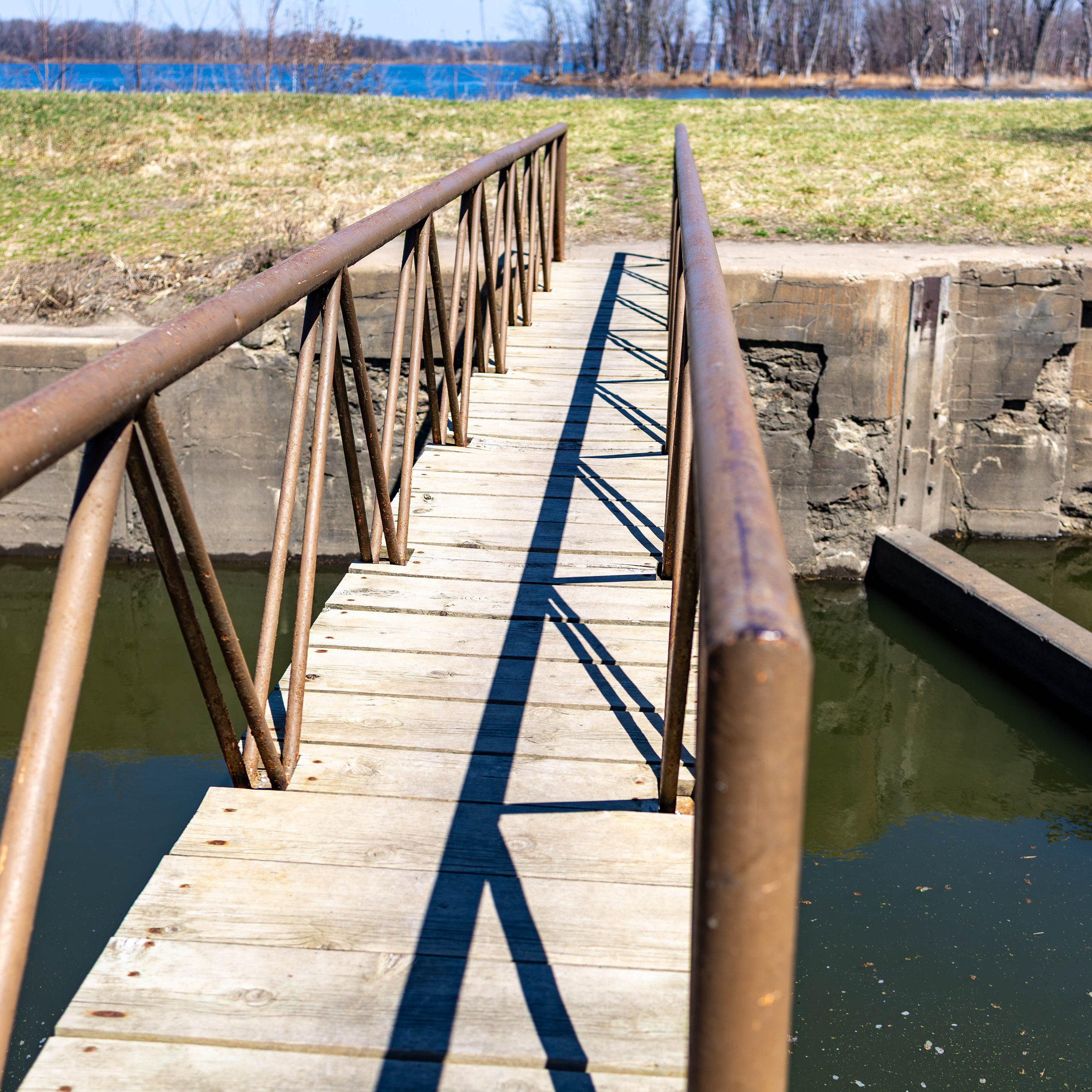 A small wooden footbridge with rusted metal railings spanning over a narrow canal or waterway, with grassy land and trees in the background under a clear sky.