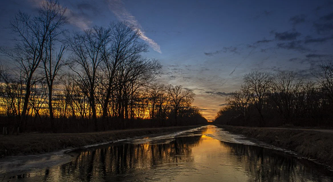 Sunset over a canal with leafless trees on both sides, reflecting the sky and trees in the water.