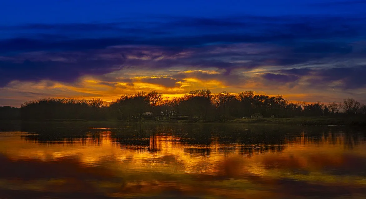Sunset over a river with trees and houses in the background, reflection on the water.