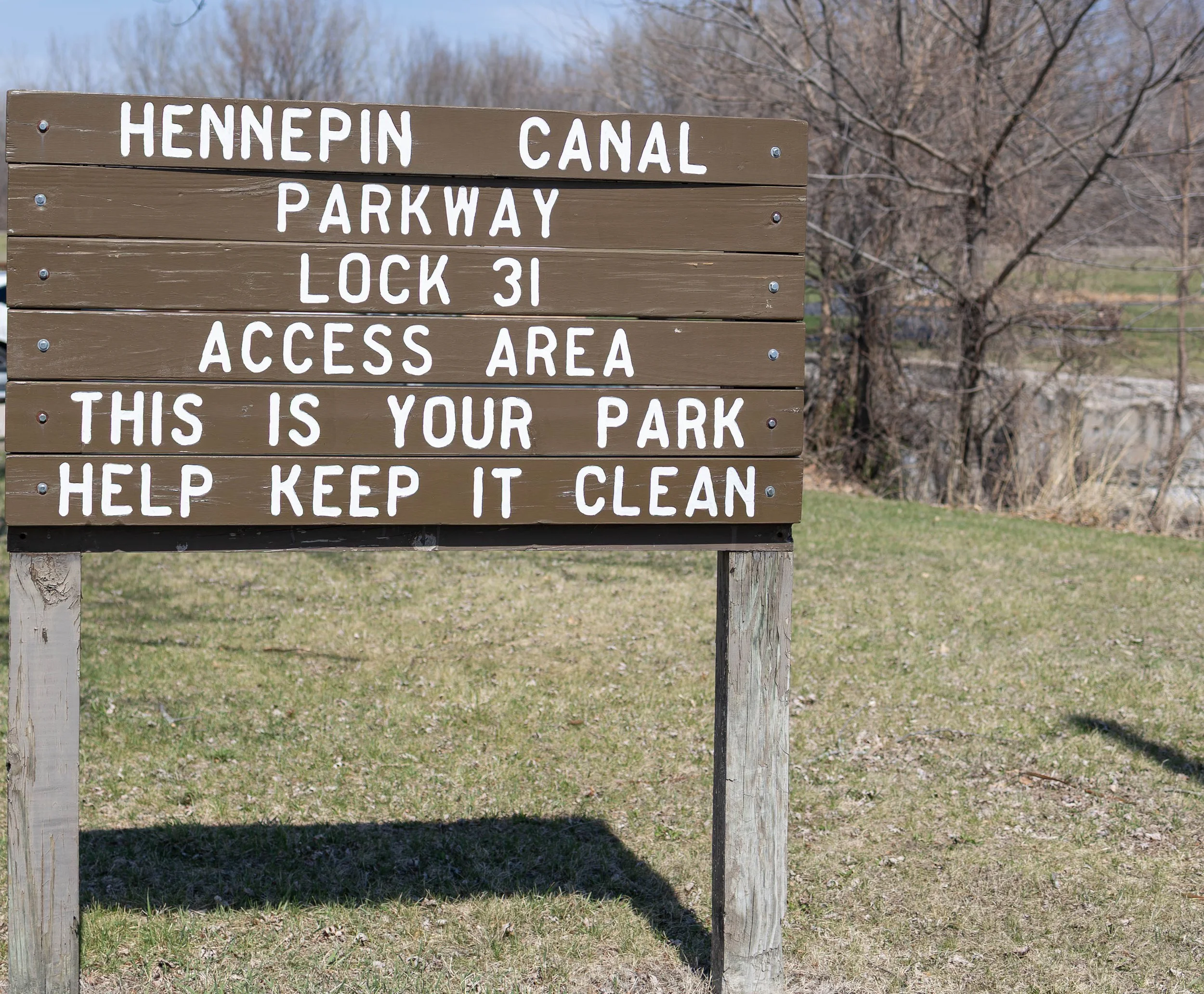 Wooden sign with white lettering that says: Hennepin Canal Parkway, Lock 31 Access Area, This is your park, Help keep it clean, in a park with grass, trees, and a river in the background.