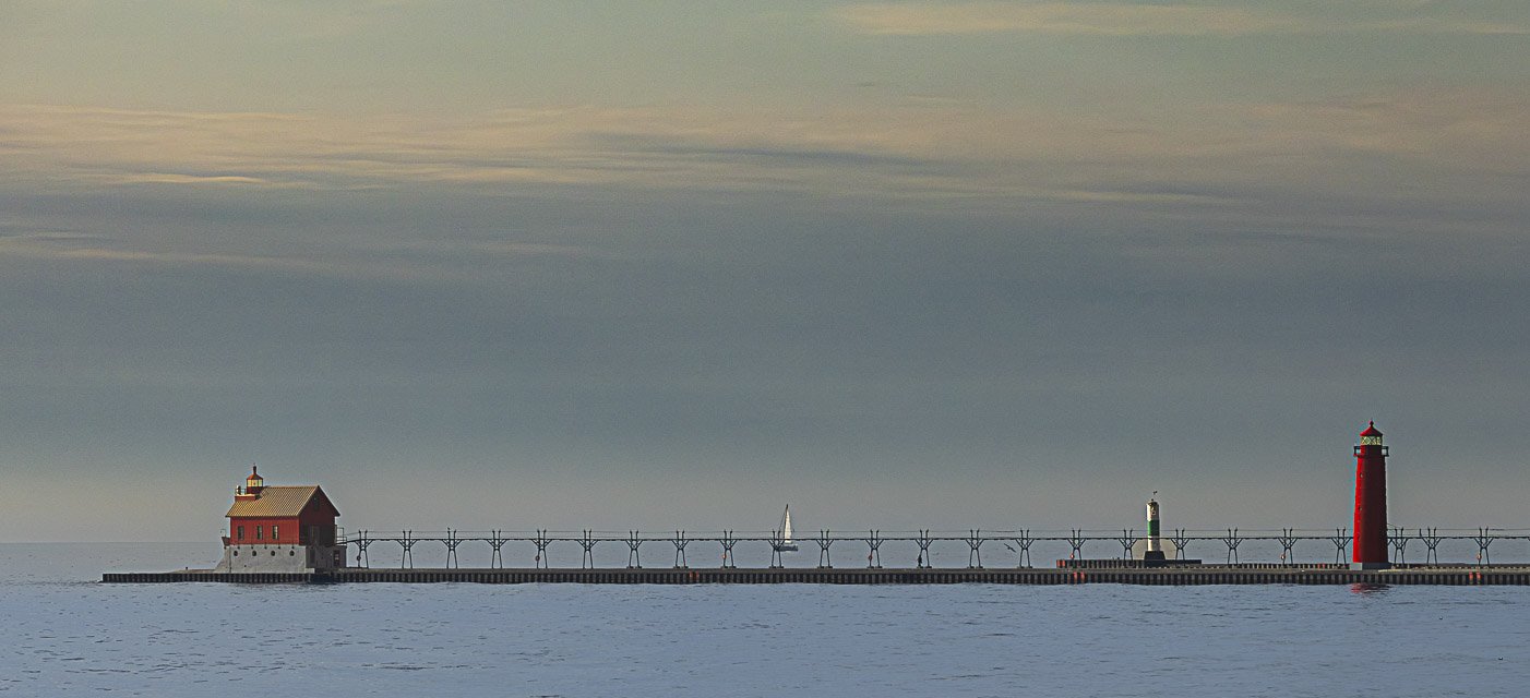Lighthouse and small building on a pier extending into a body of water with a sailboat in the background.