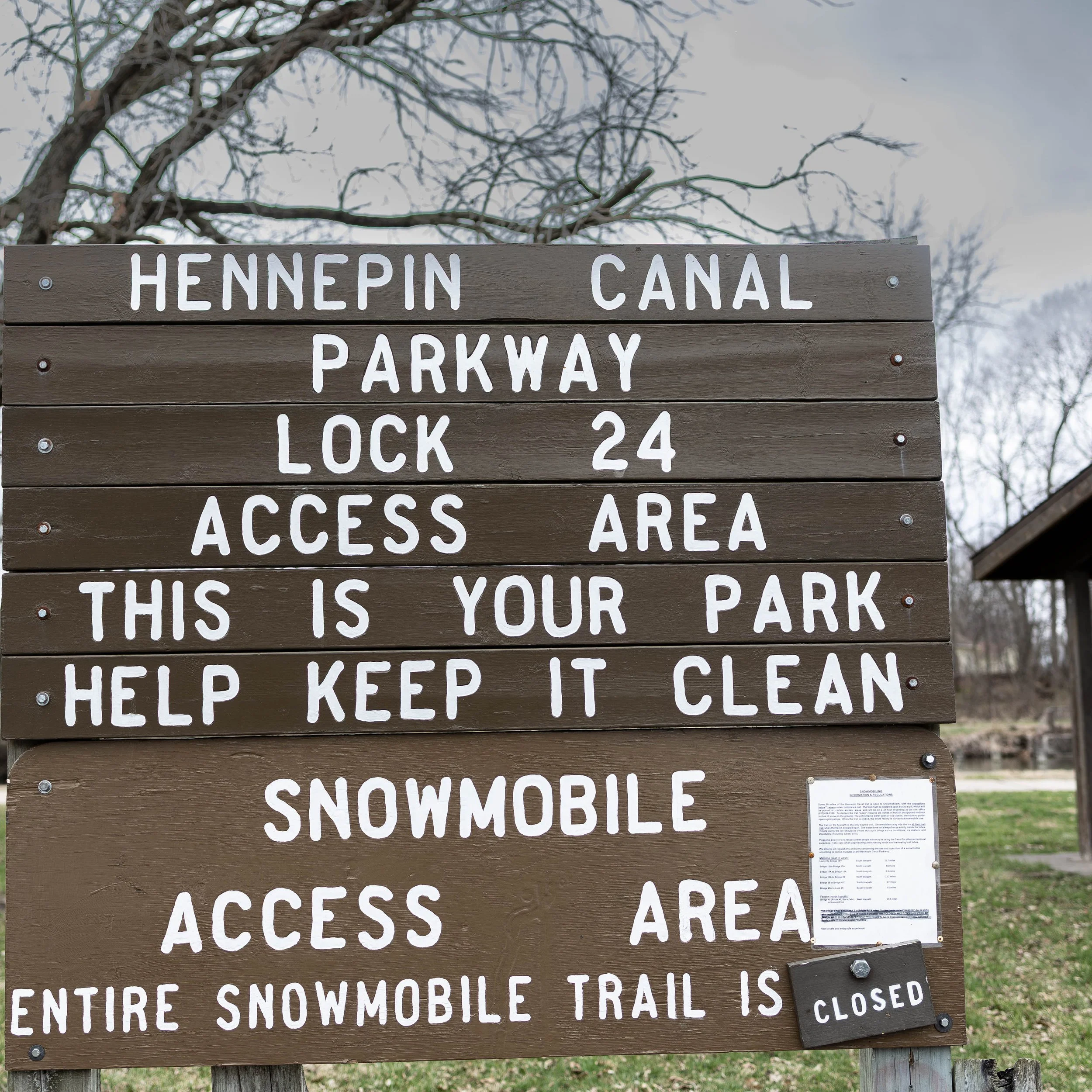 Brown informational signboard with white text about Hennepin Canal Parkway Lock 24 access area and snowmobile trail closure, outdoors with leafless trees and cloudy sky in the background.