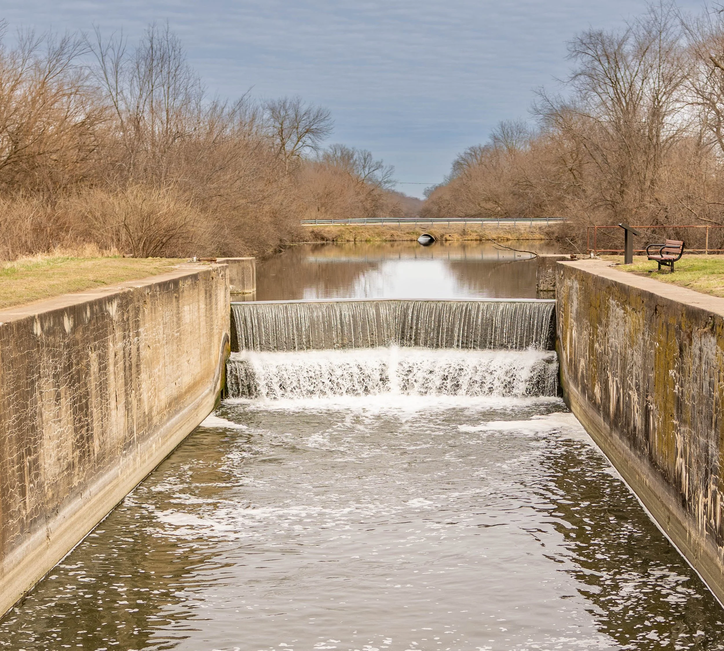 A small dam on a river, with water flowing over a concrete ledge, surrounded by leafless trees and a park bench on the right side.