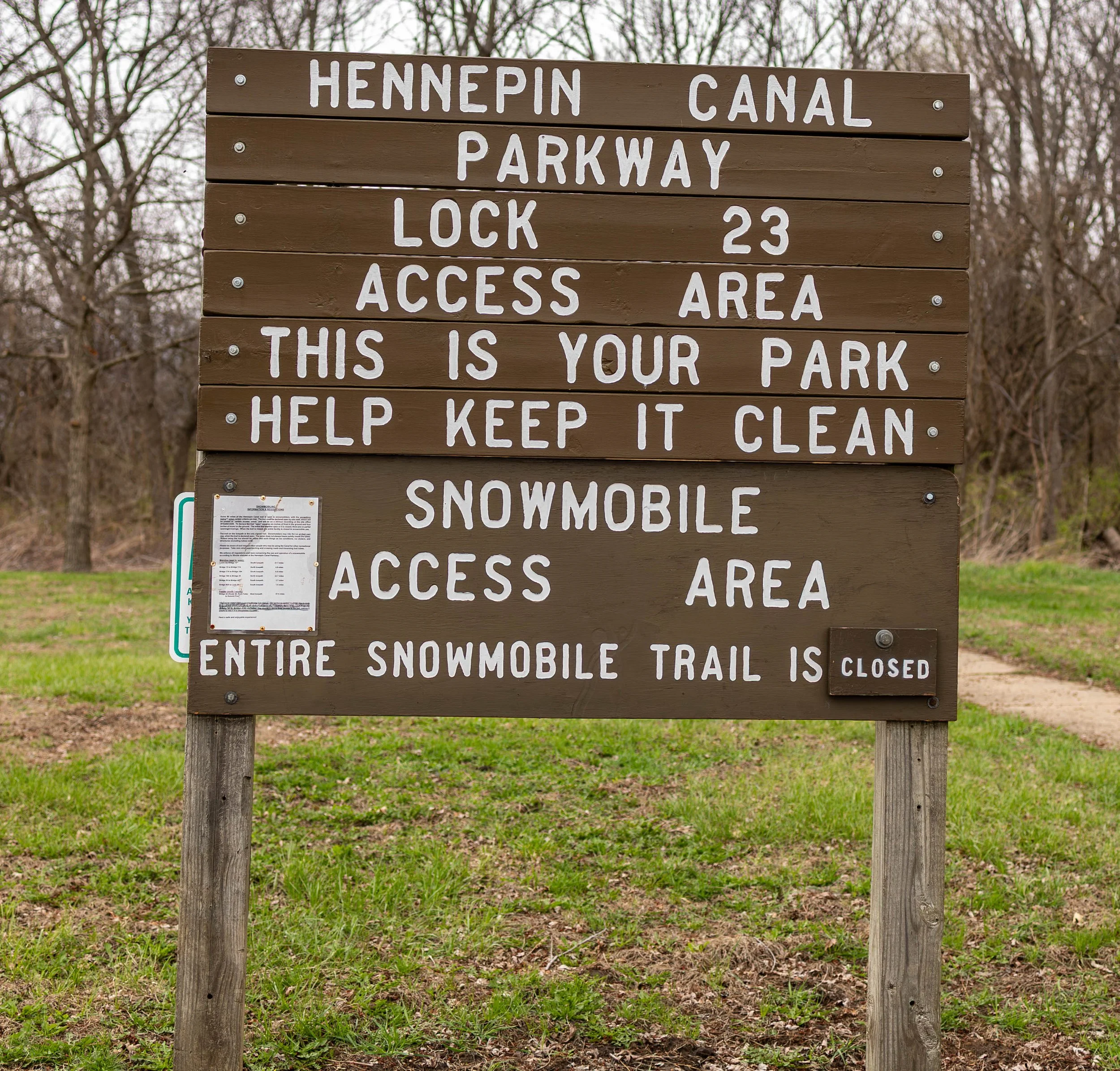 Wooden park sign with white lettering informing about Hennepin Canal Parkway Lock 23 and snowmobile trail closure, urging visitors to keep the park clean.