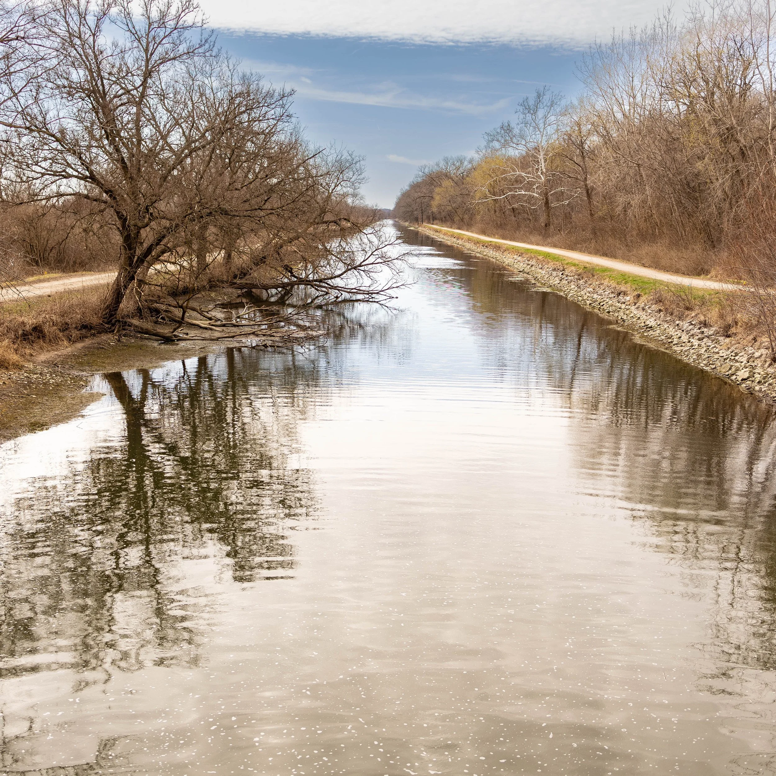 Calm waterway with reflections, trees on the banks, clear sky, bare trees indicating early spring or late fall, with a dirt pathway running alongside the canal.