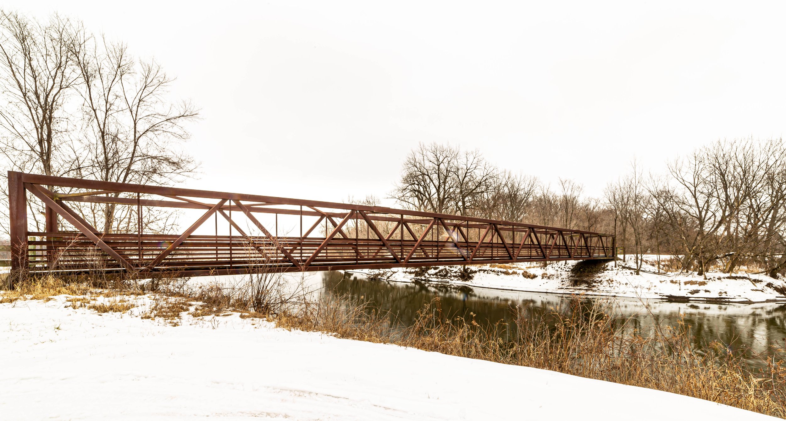 A snowy landscape with a brown metal footbridge crossing a river, bare trees in the background under an overcast sky.