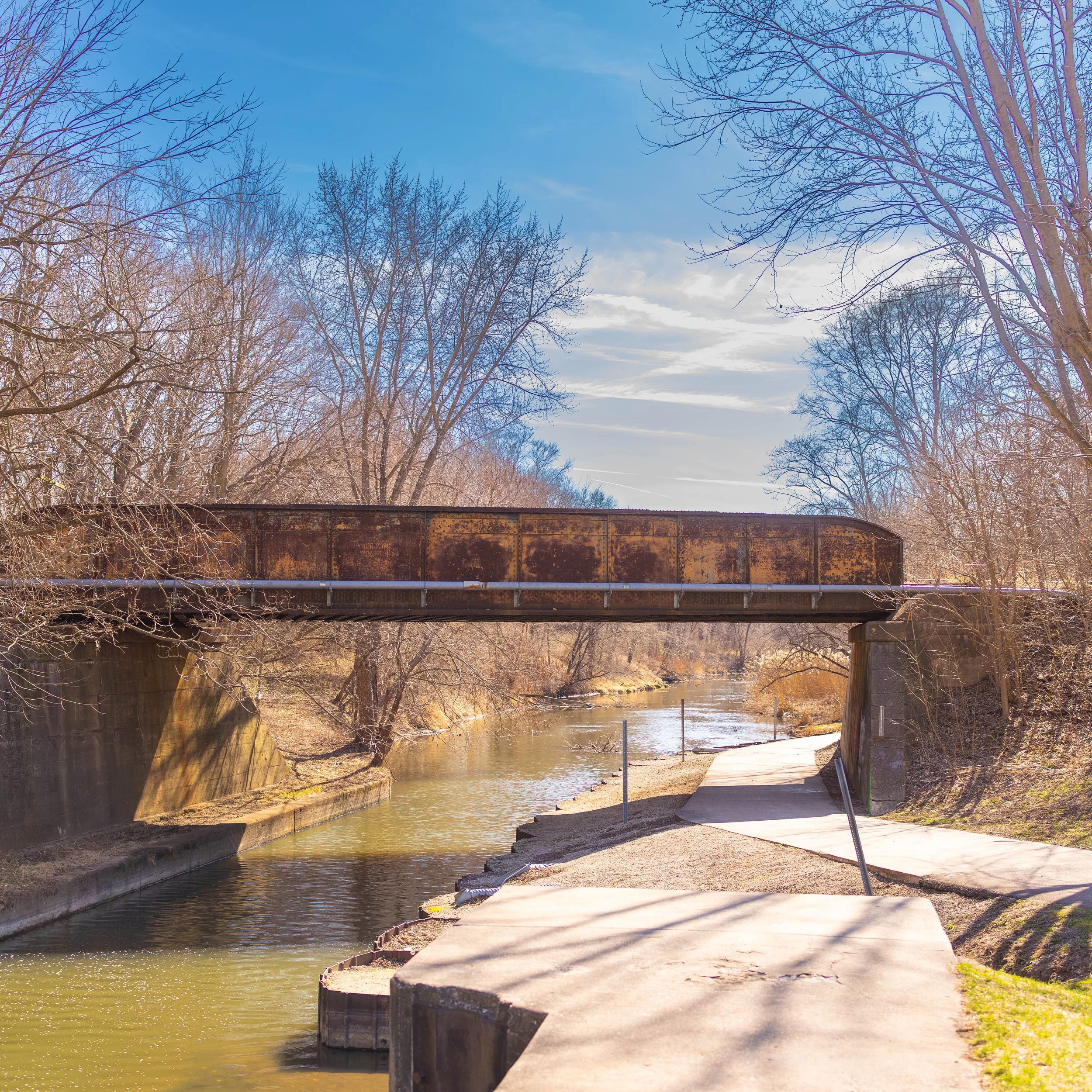 A rusty train bridge spans over a narrow river, with leafless trees along the riverbank and a paved walkway beside the water on a sunny day with a partly cloudy sky.