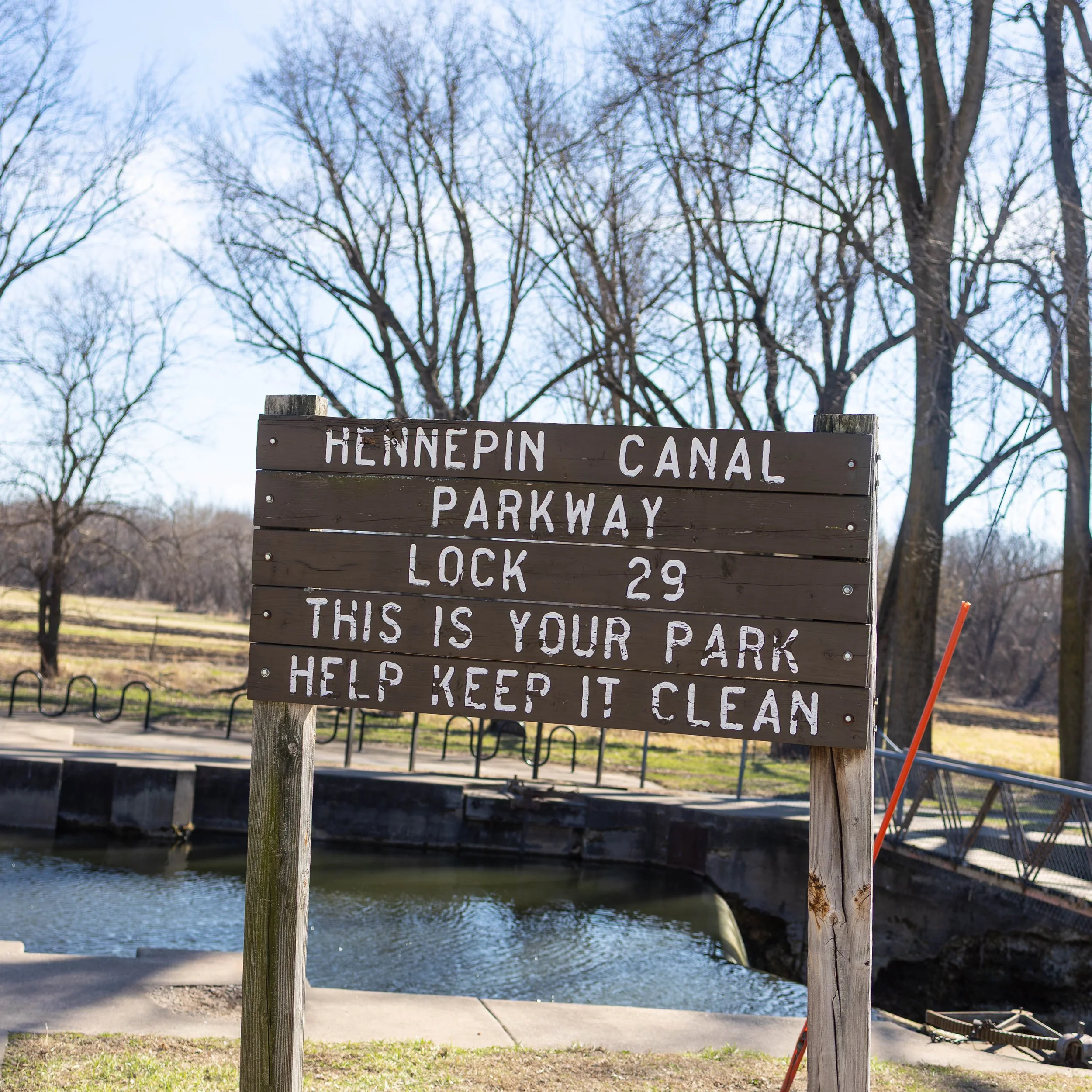 A wooden sign near water in a park reads: 'Hennepin Canal Parkway Lock 29. This is your park. Help keep it clean.' in front of trees with bare branches during daytime.