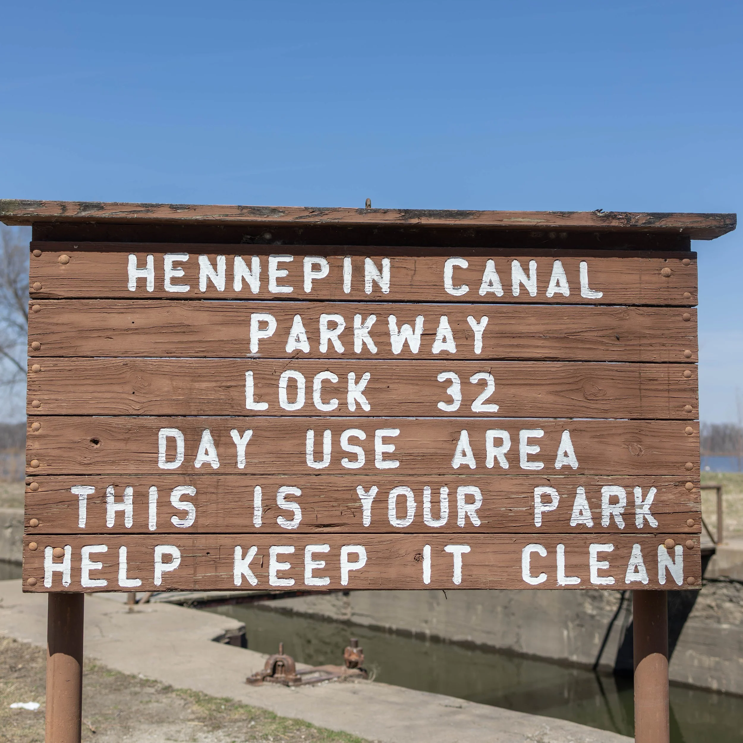 A wooden sign with white painted text that reads: "HENNEPIN CANAL PARKWAY LOCK 32 DAY USE AREA THIS IS YOUR PARK HELP KEEP IT CLEAN." The sign is placed outdoors with a blue sky and a canal in the background.