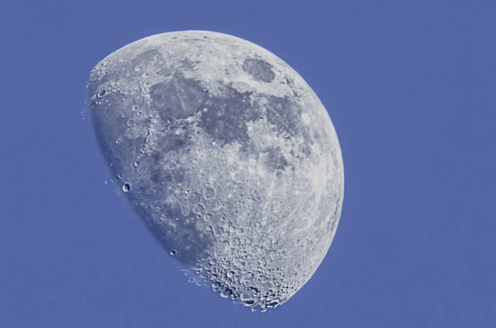 A close-up photograph of the moon showing detailed craters against a blue sky.