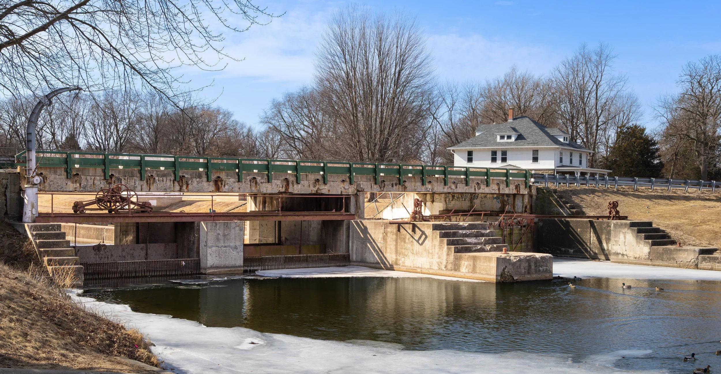 A canal lock with a green and rust-colored metal gate, an old hogshead wheel mechanism, concrete walls, stairs, and steps leading into the water, with a house and leafless trees in the background on a clear day.