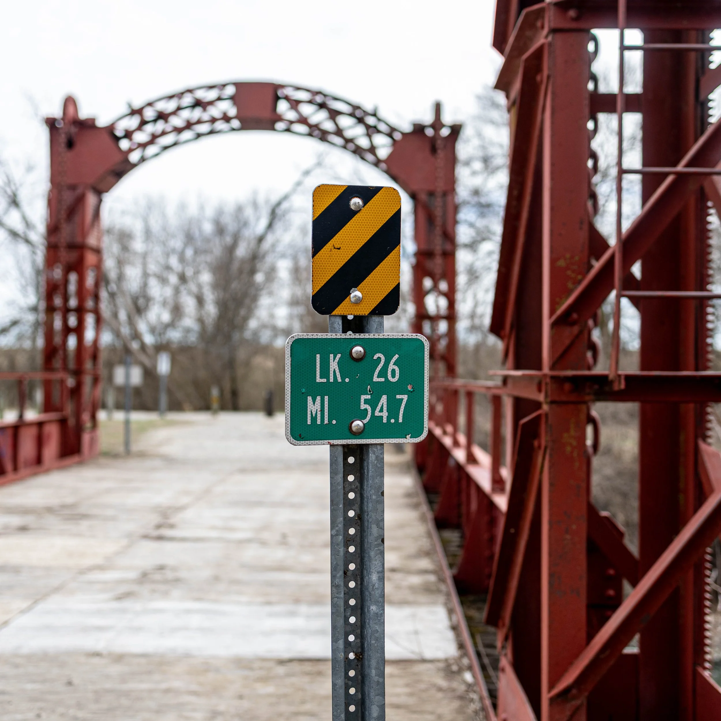 Close-up of a roadside signpost with a black and yellow striped warning sign on top and a green informational sign below, indicating mile markers for Lake (LK.) 26 and 54.7 miles.