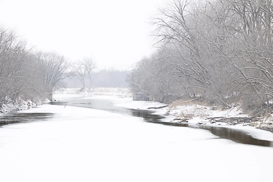 Snow-covered river with leafless trees on both sides, overcast sky, and patches of ice on the water.
