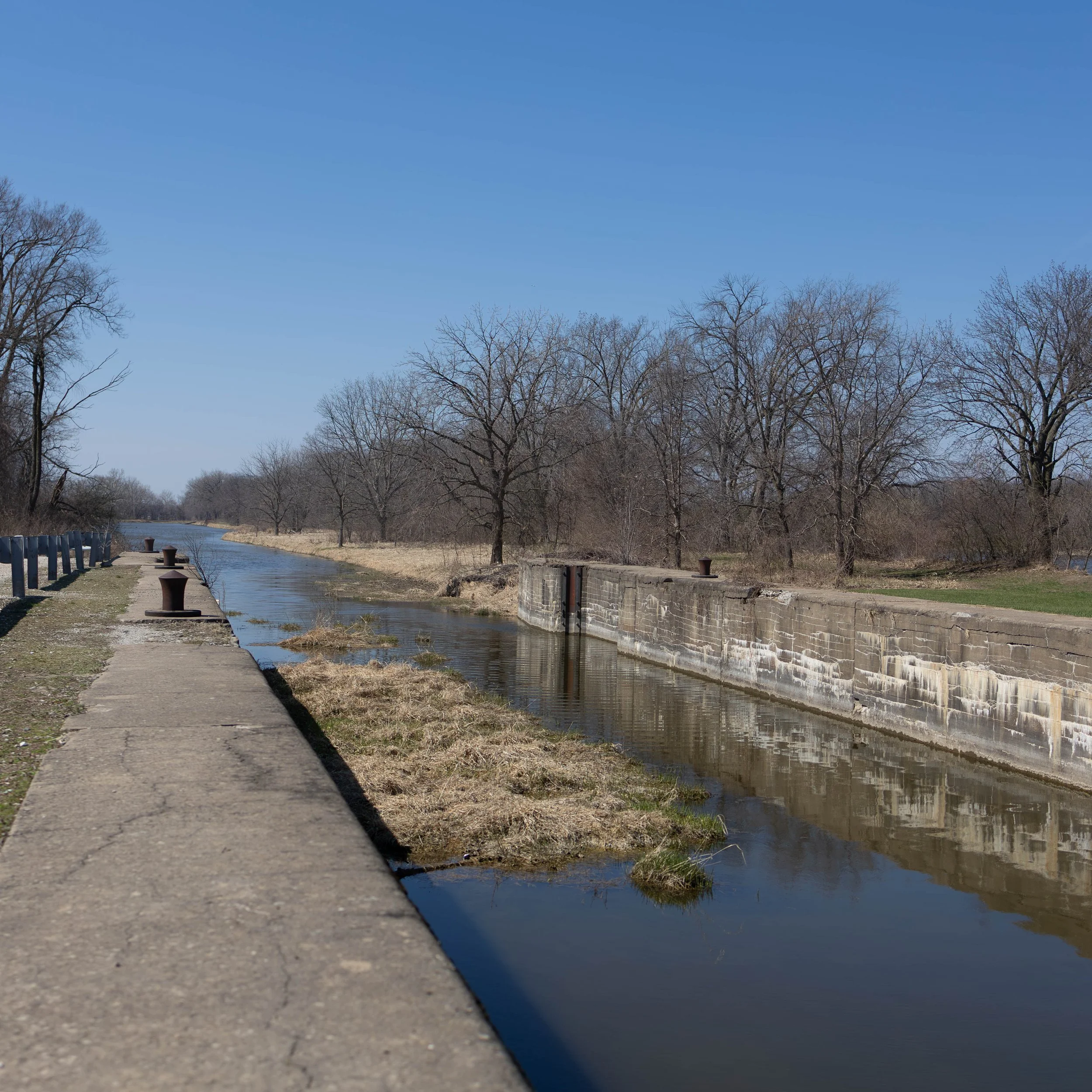 A walkway beside a canal with leafless trees and a blue sky in the background.