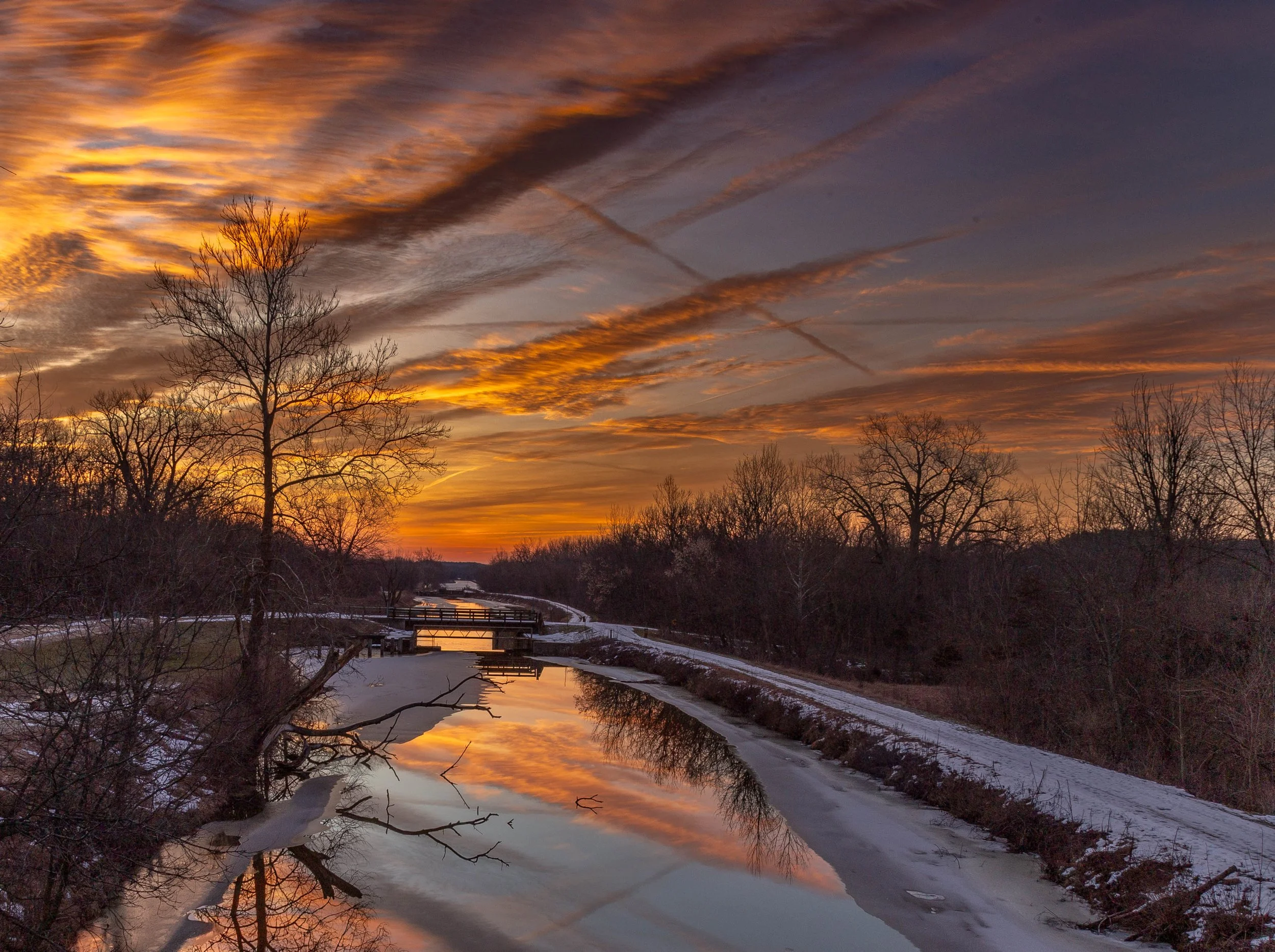 Sunset over a partially frozen river with leafless trees on both sides, a small bridge in the distance, and colorful sky with clouds reflecting on the water.