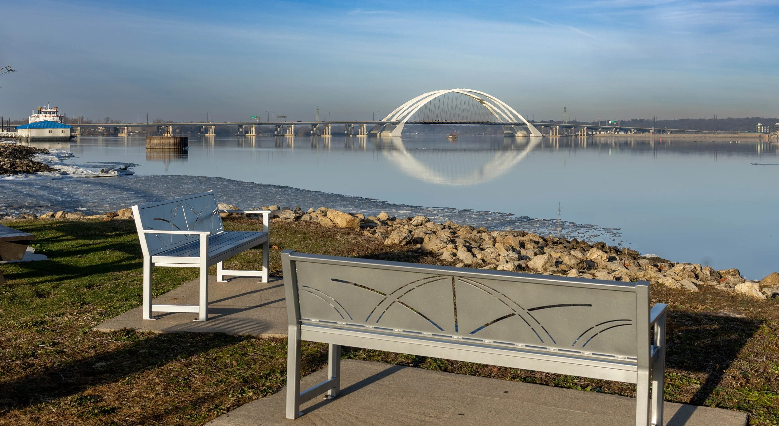 A peaceful riverside scene featuring a wide, calm river with a modern white arch bridge reflecting on the water. In the foreground, two empty benches are positioned on a paved area beside some rocks and grass, with a clear sky overhead.