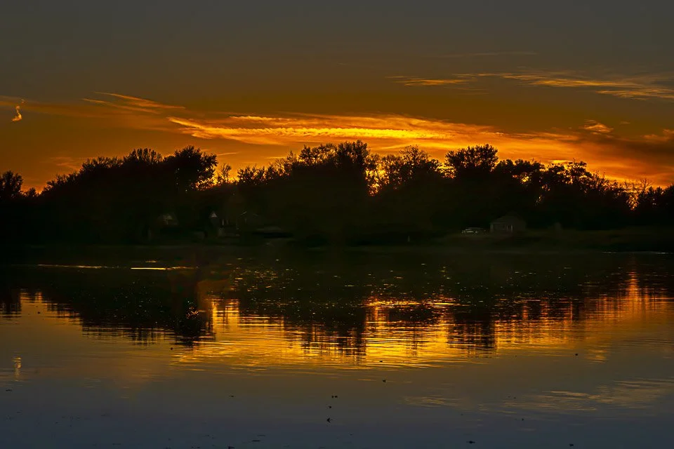 Sunset over a lake with silhouetted trees and houses, orange and yellow clouds reflected in the water.