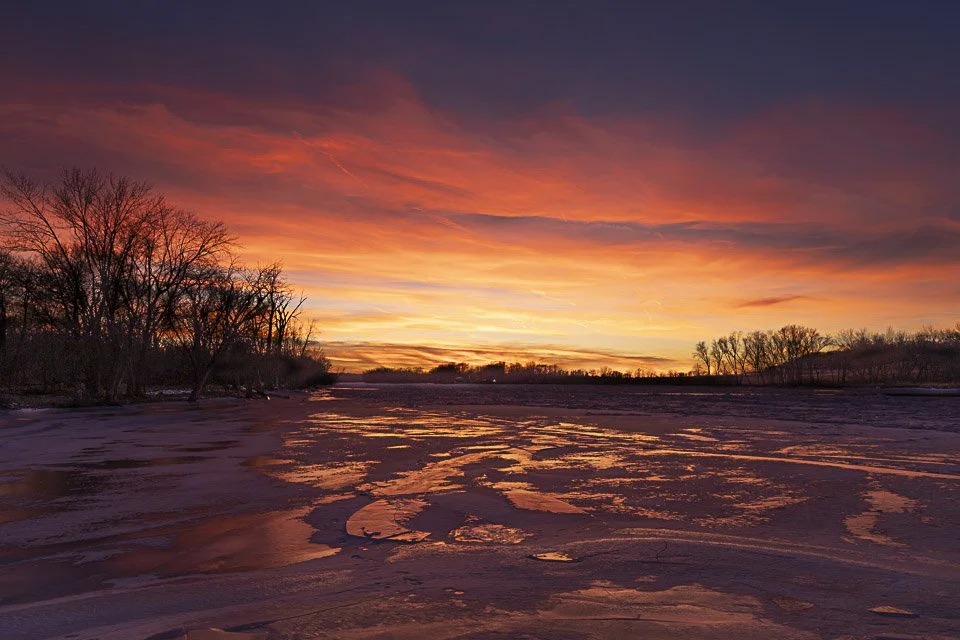 A colorful sunset over a frozen river with bare trees along the shoreline.