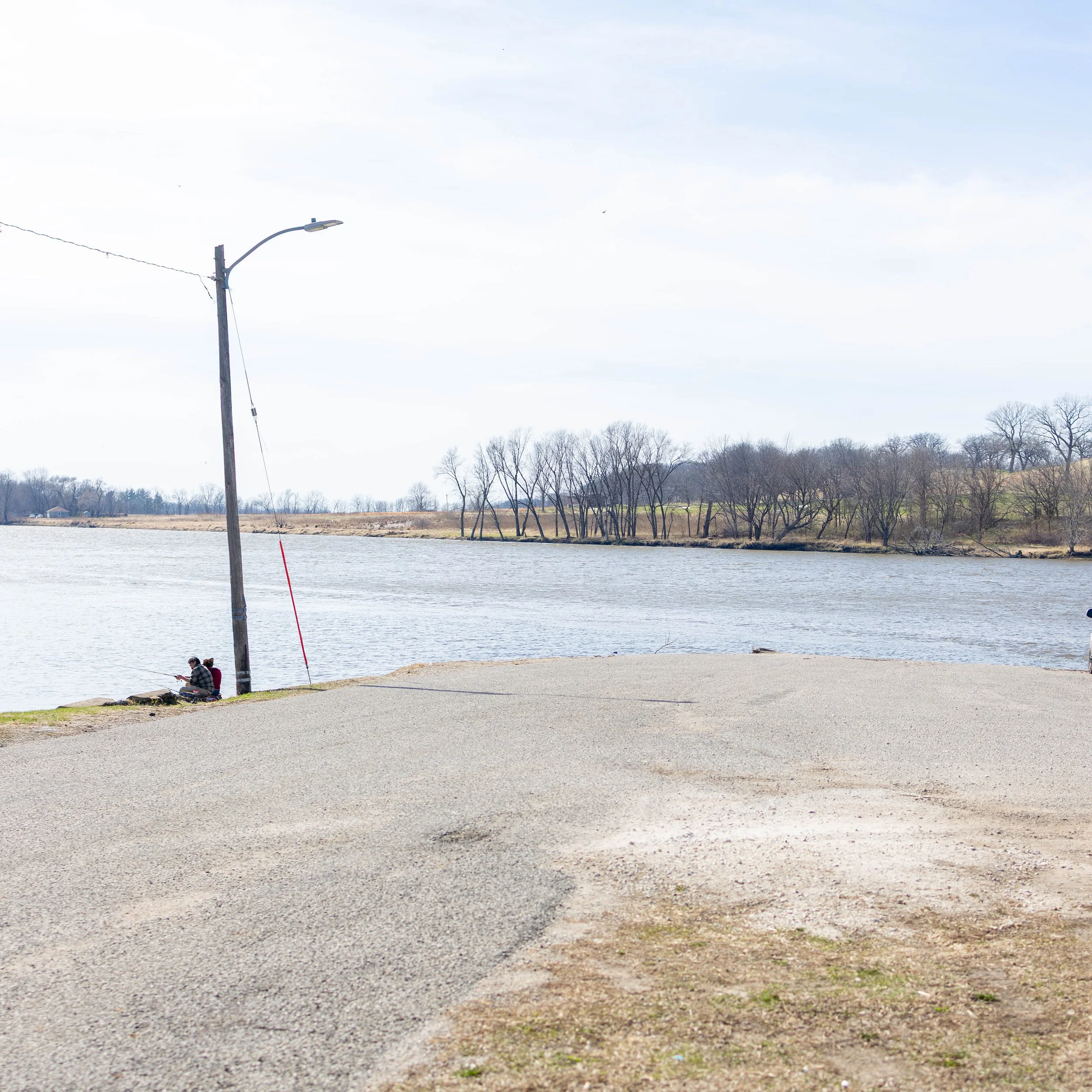 Two people sitting on rocks near a body of water, with a fishing pole propped against a street lamp, and trees in the background during daytime.