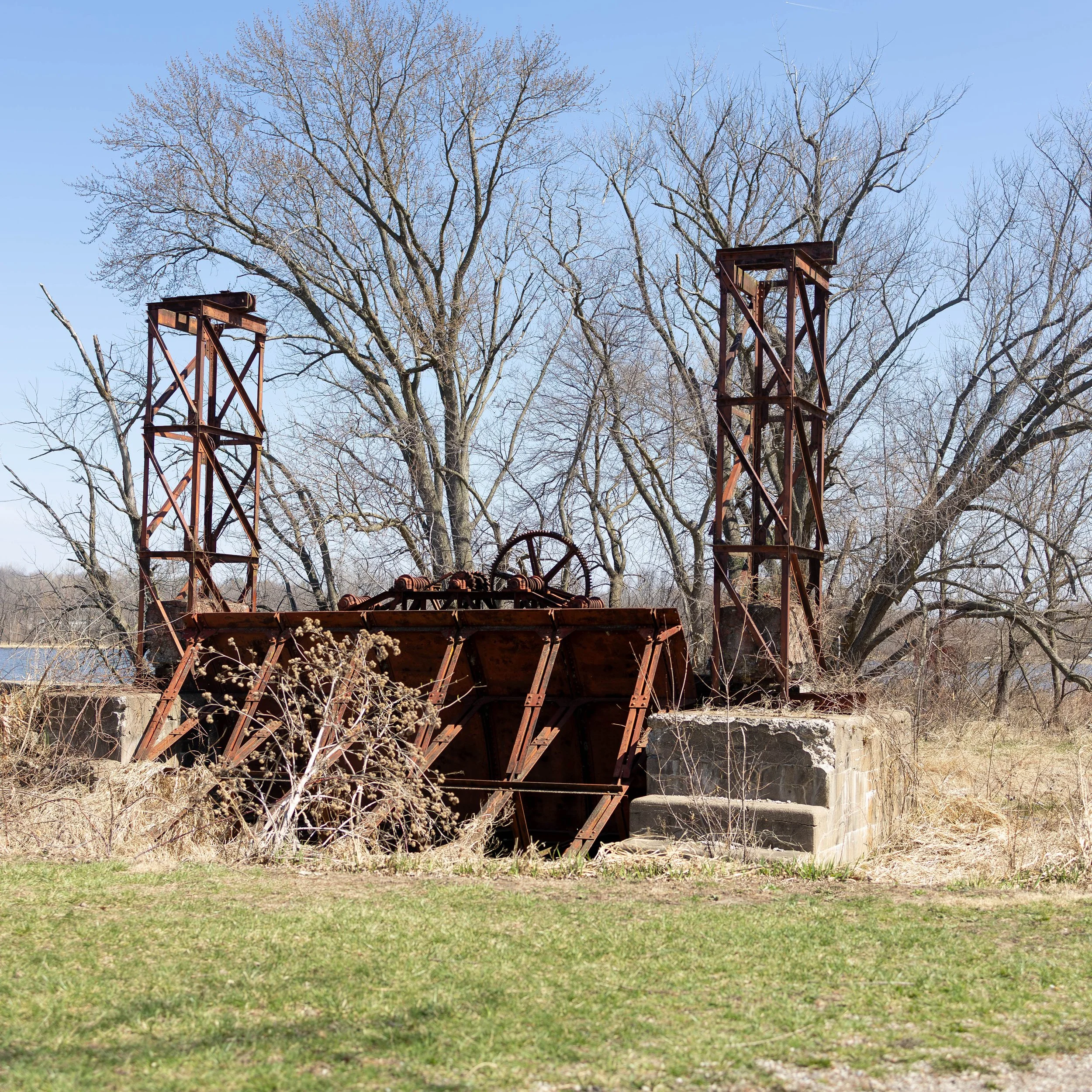 Old, rusted industrial equipment outdoors with leafless trees in background and a river on the left side