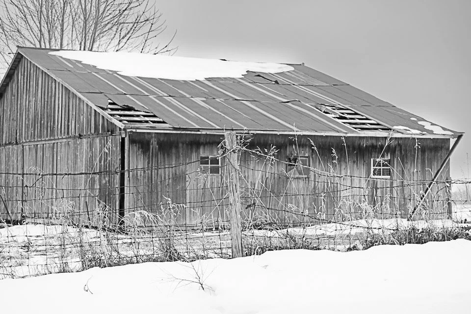 An old wooden barn with a partially damaged roof in a snow-covered landscape.