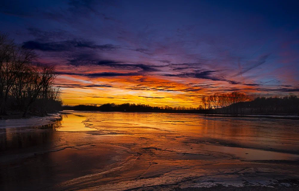 A sunset over a river with reflected orange, pink, and purple colors in the sky and water, with leafless trees along the shoreline.