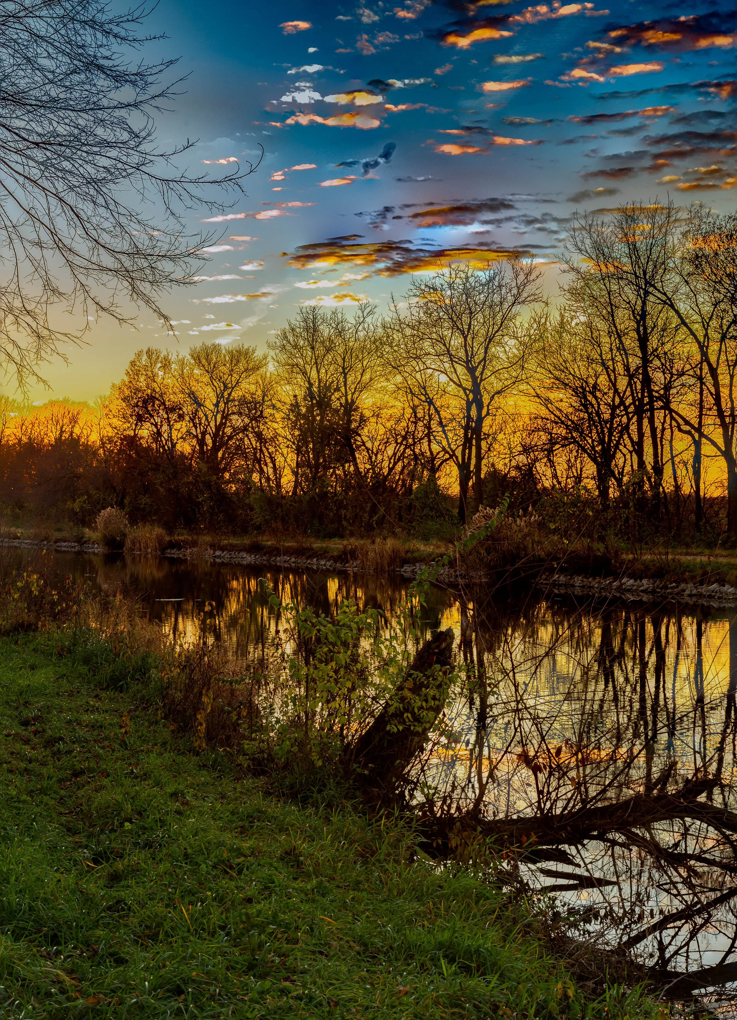 Sunset over a calm river with bare trees on the bank, reflected in the water, and a partly cloudy sky with warm colors.