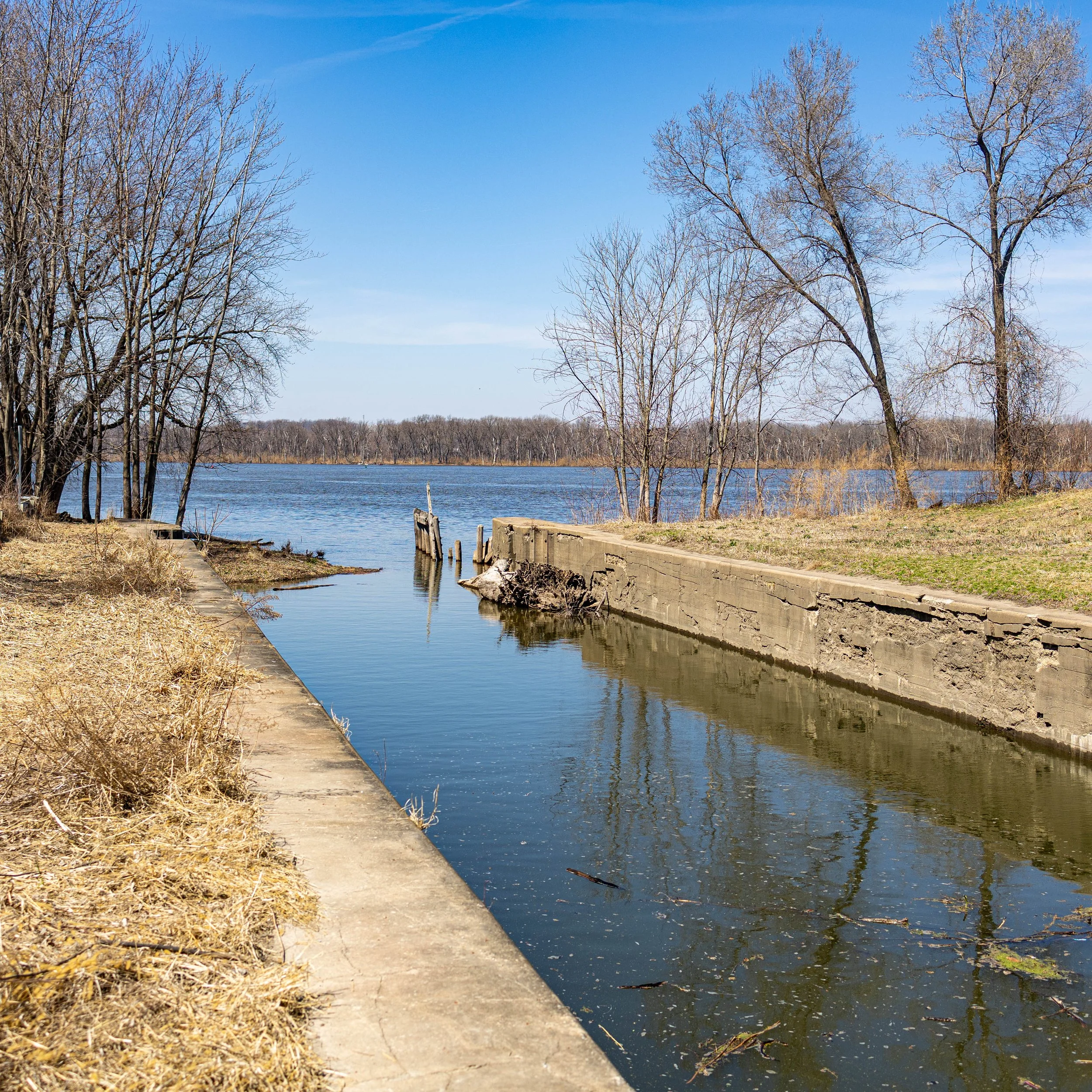 A small canal or creek with a concrete bank on the right and a grassy area on the left, lined with leafless trees and a body of water stretching into the distance on a clear day.