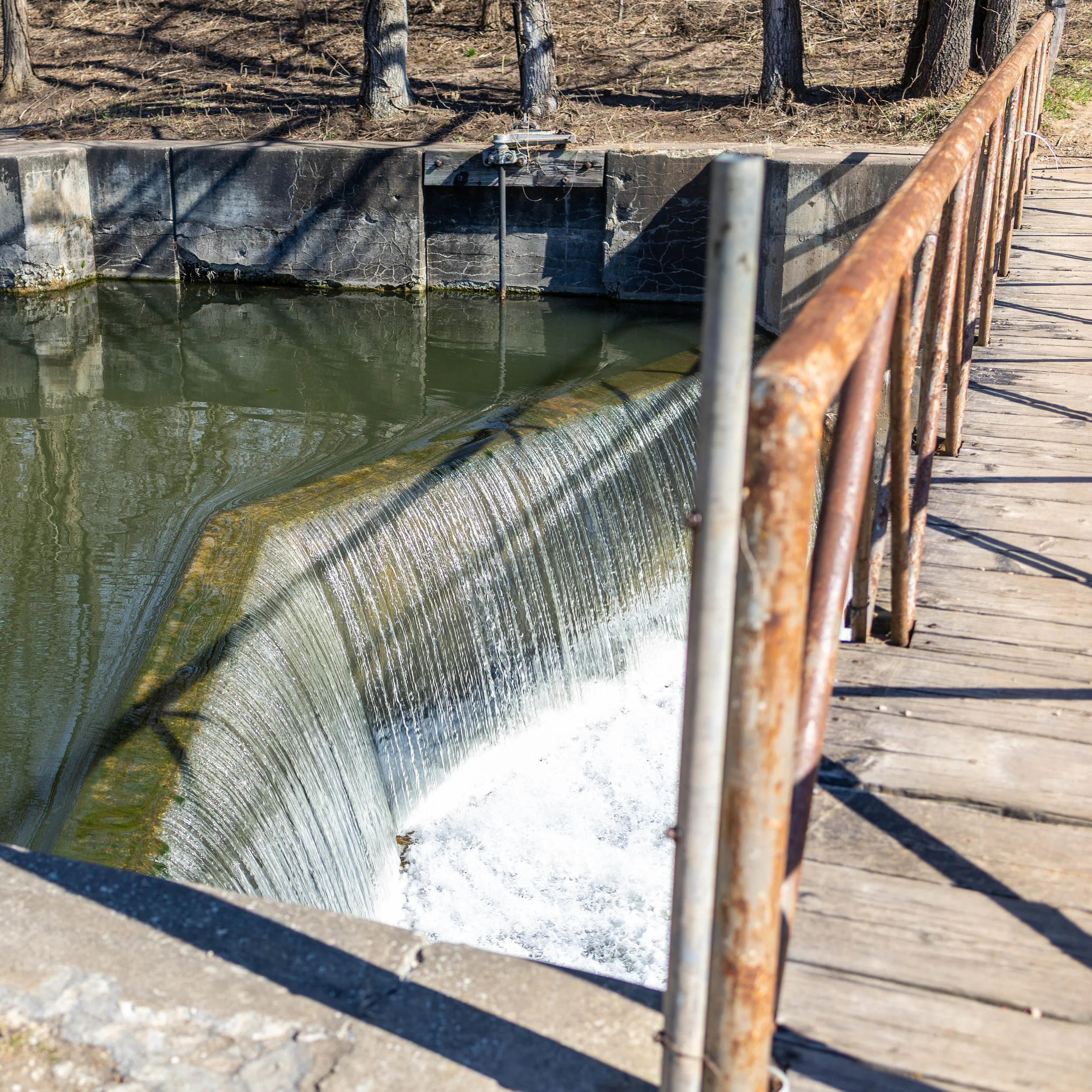 A small dam or weir in a river with water flowing over it, surrounded by a rusty metal railing and a wooden walkway.