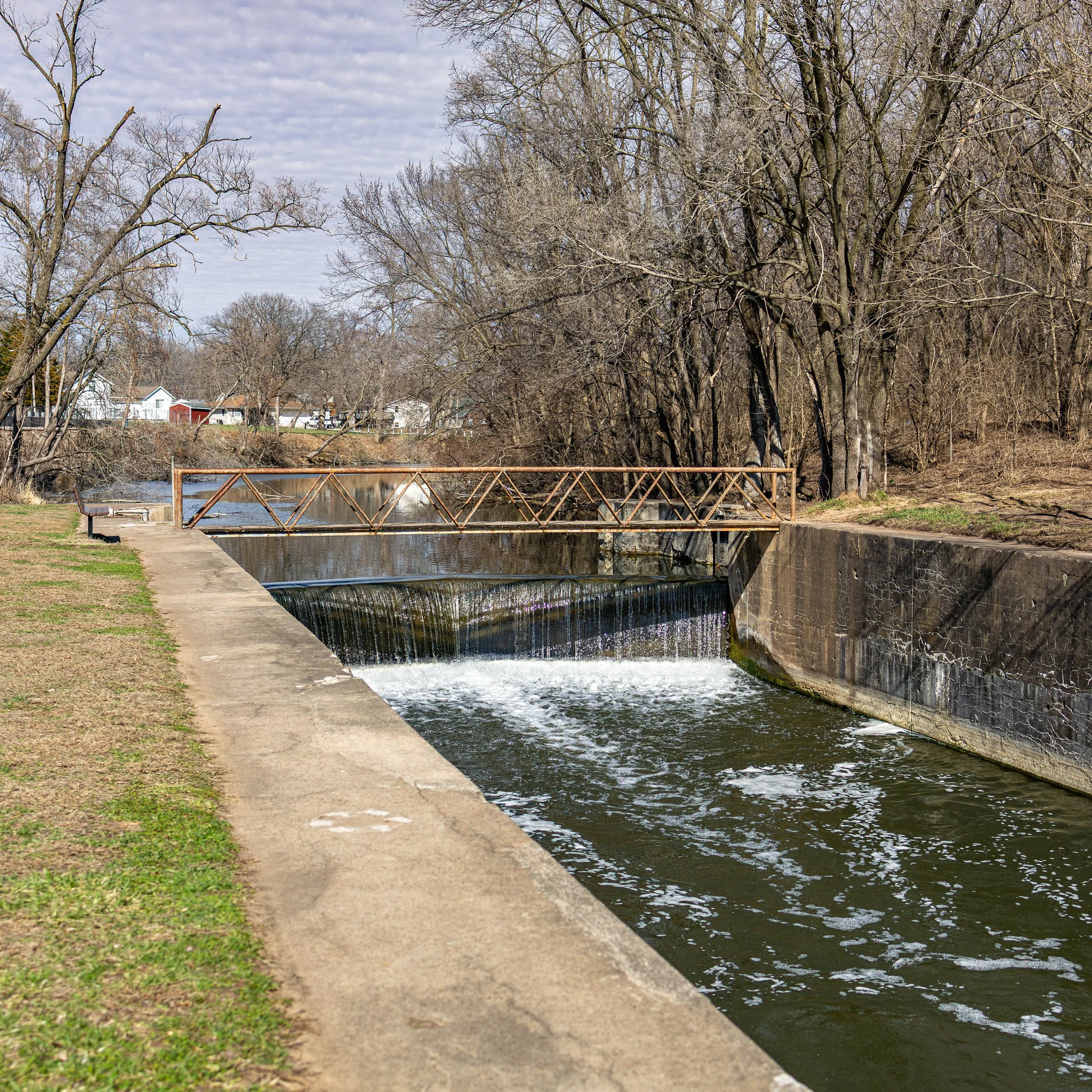 Scene of a small waterway with a concrete wall, a metal footbridge, leafless trees, and houses in the background during overcast weather.