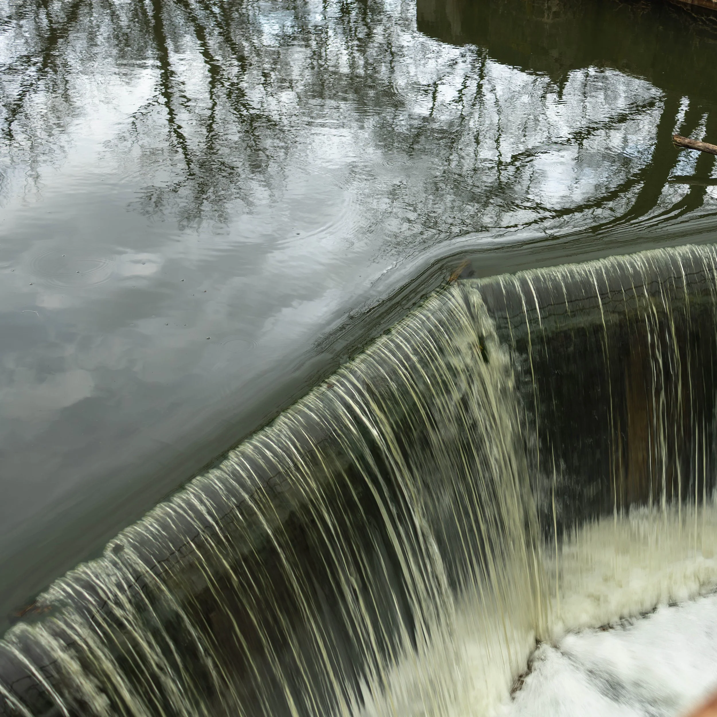 A close-up of a waterfall over a curved concrete structure, with water flowing down and trees reflected in the water surface above.