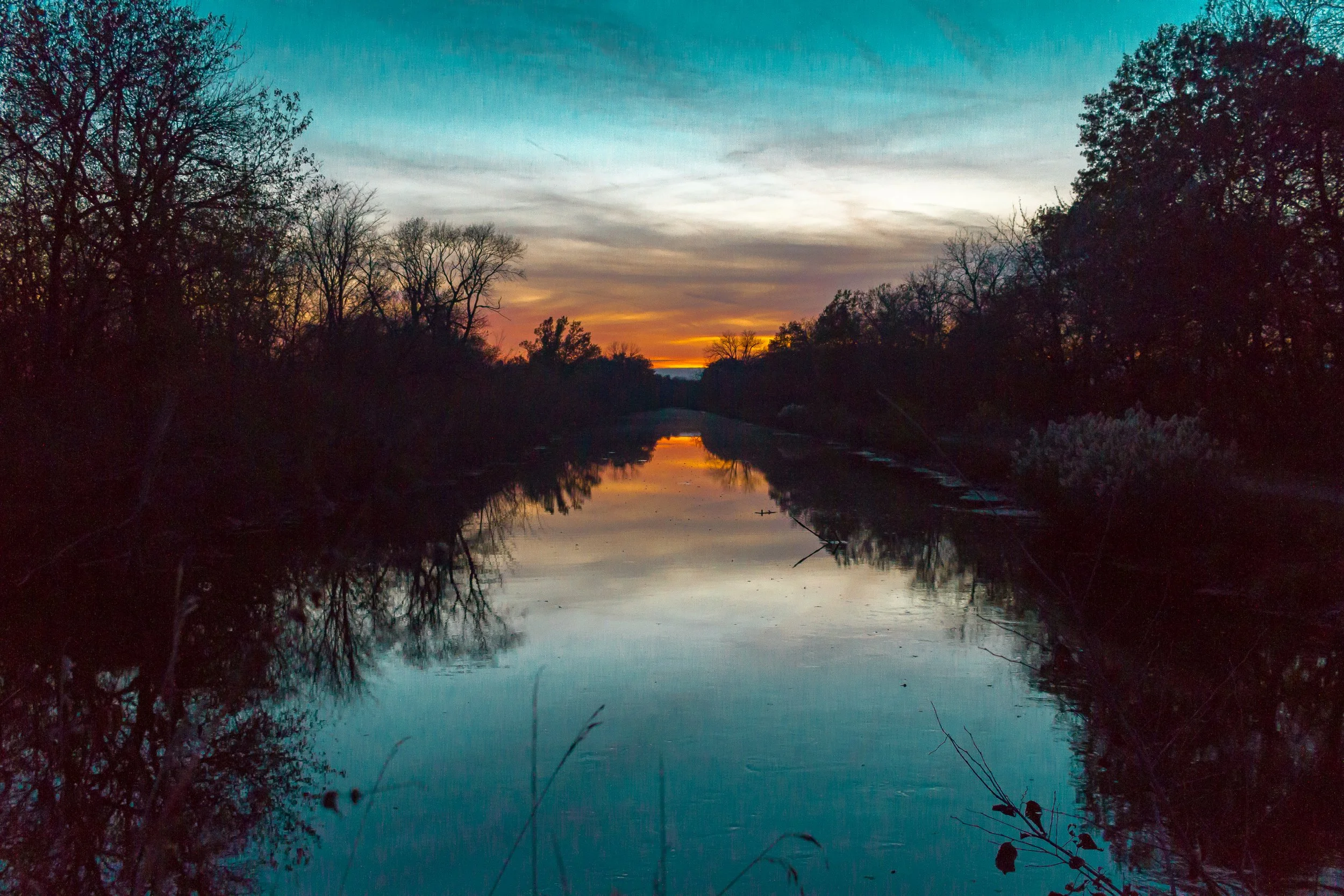 A peaceful river at sunset with trees on both sides reflecting in the water and a colorful sky.