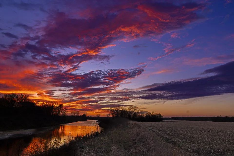 A vibrant sunset over a river with colorful clouds in shades of purple, pink, orange, and red, silhouetted trees along the riverbank, and fields in the foreground.