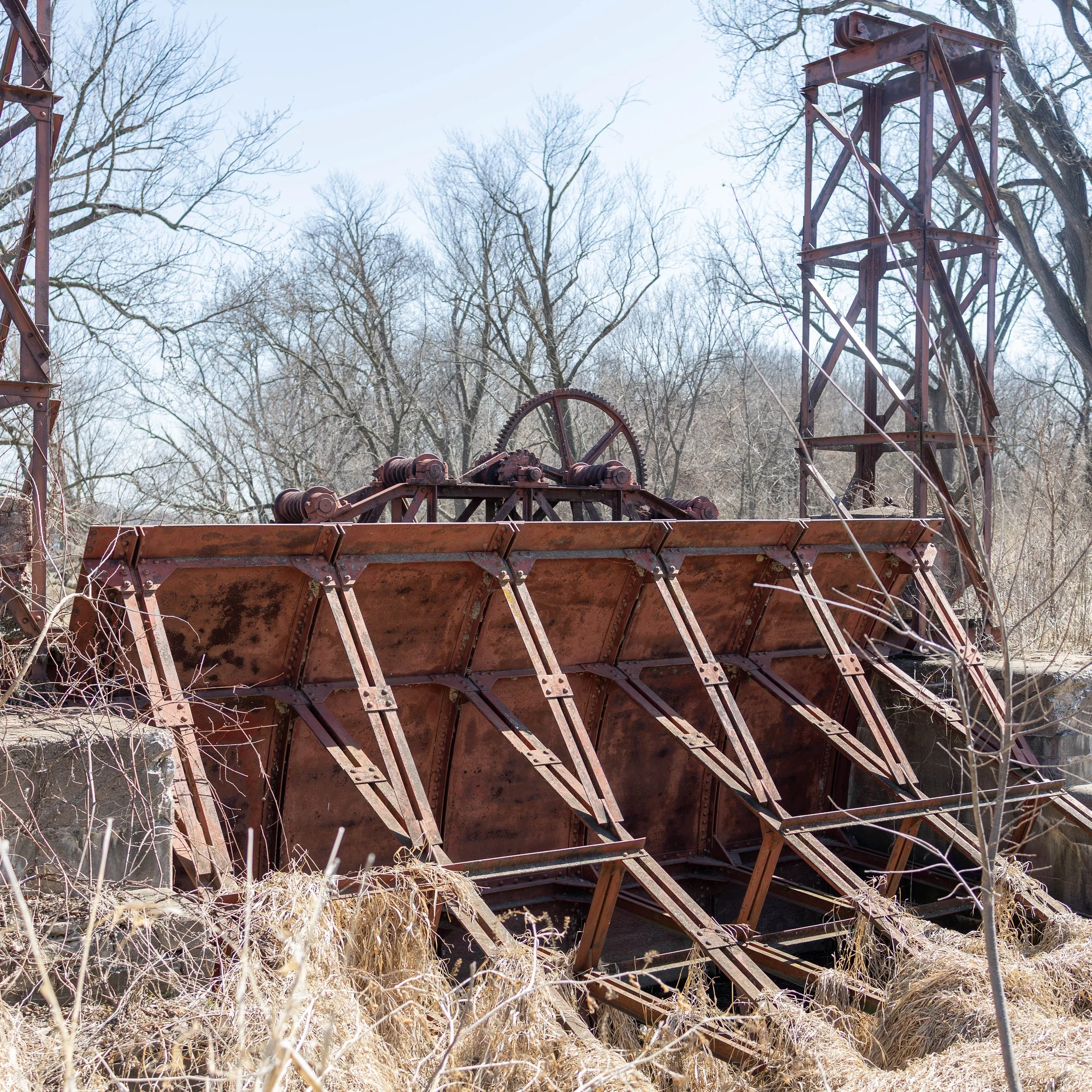 Old rusty industrial waterwheel structure outdoors among leafless trees and dry grass in early spring.