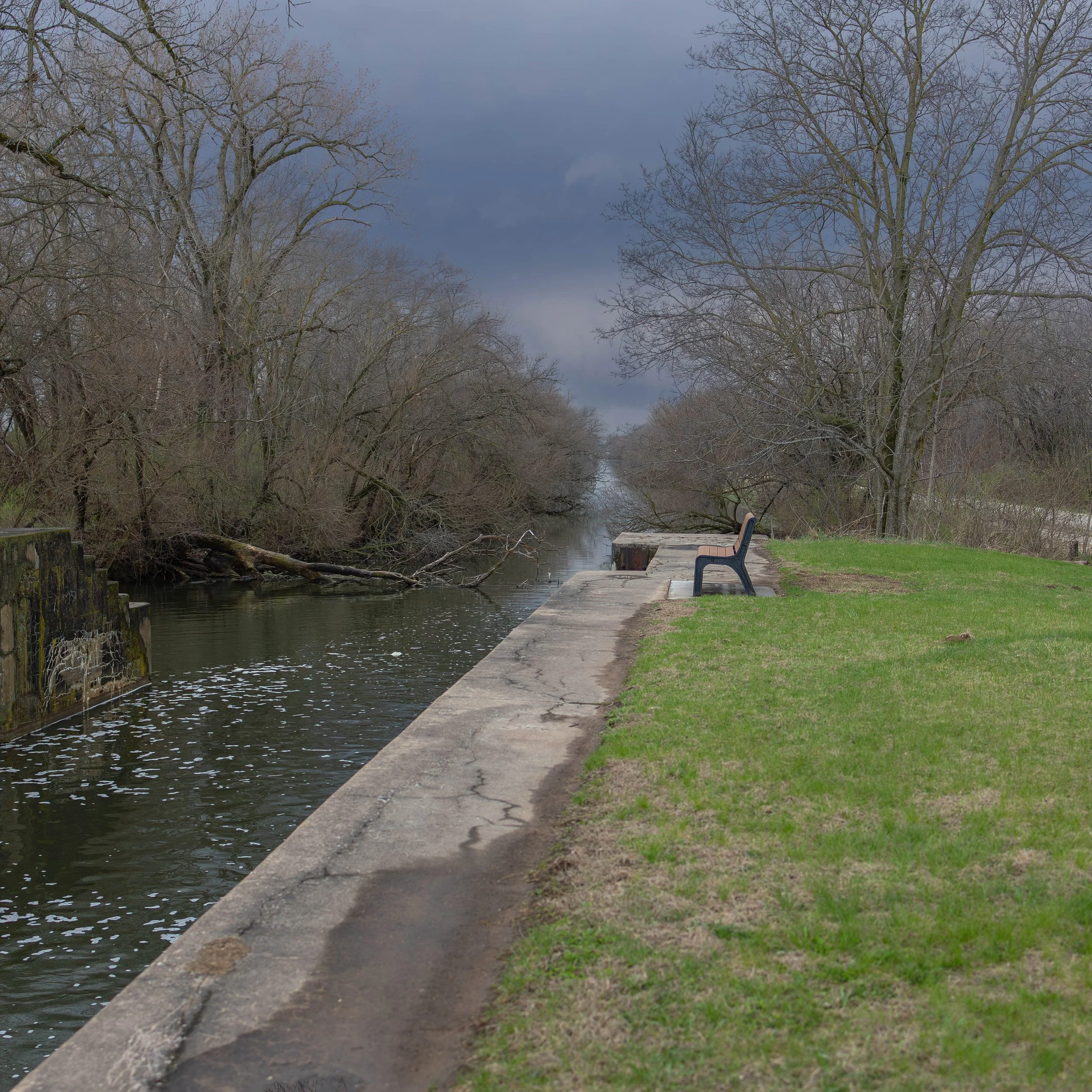 A park with a narrow waterway on the left, lined by leafless trees. There's a grassy area with a bench facing the water on the right. The sky is overcast with dark clouds.