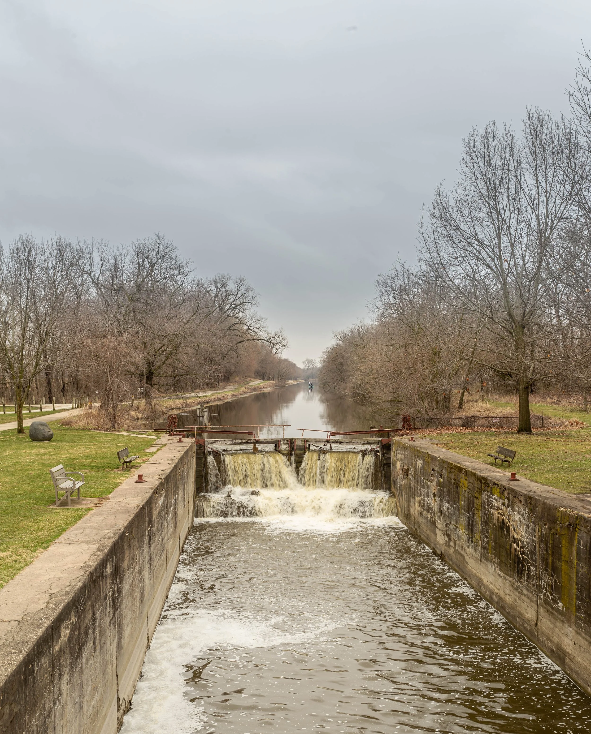 A canal with a small waterfall in a park setting, surrounded by leafless trees and benches on each side, under a cloudy sky.