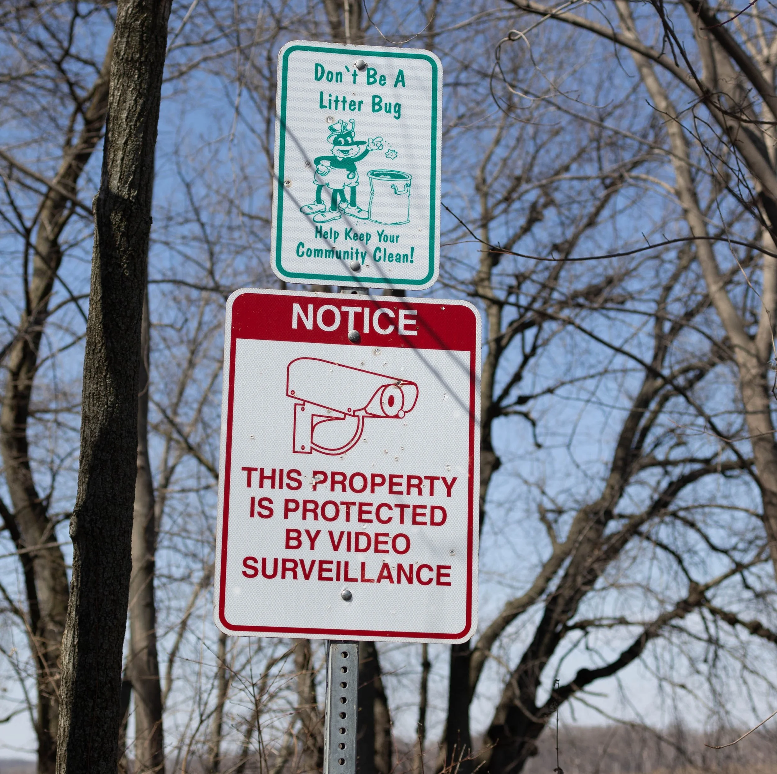 Two signs on a pole against a backdrop of leafless trees and blue sky. The upper sign is green and white, cartoon character with a trash can, saying, "Don't Be A Litter Bug. Help Keep Your Community Clean!" The lower sign is red and white, with a camera icon, stating, "NOTICE: THIS PROPERTY IS PROTECTED BY VIDEO SURVEILLANCE."