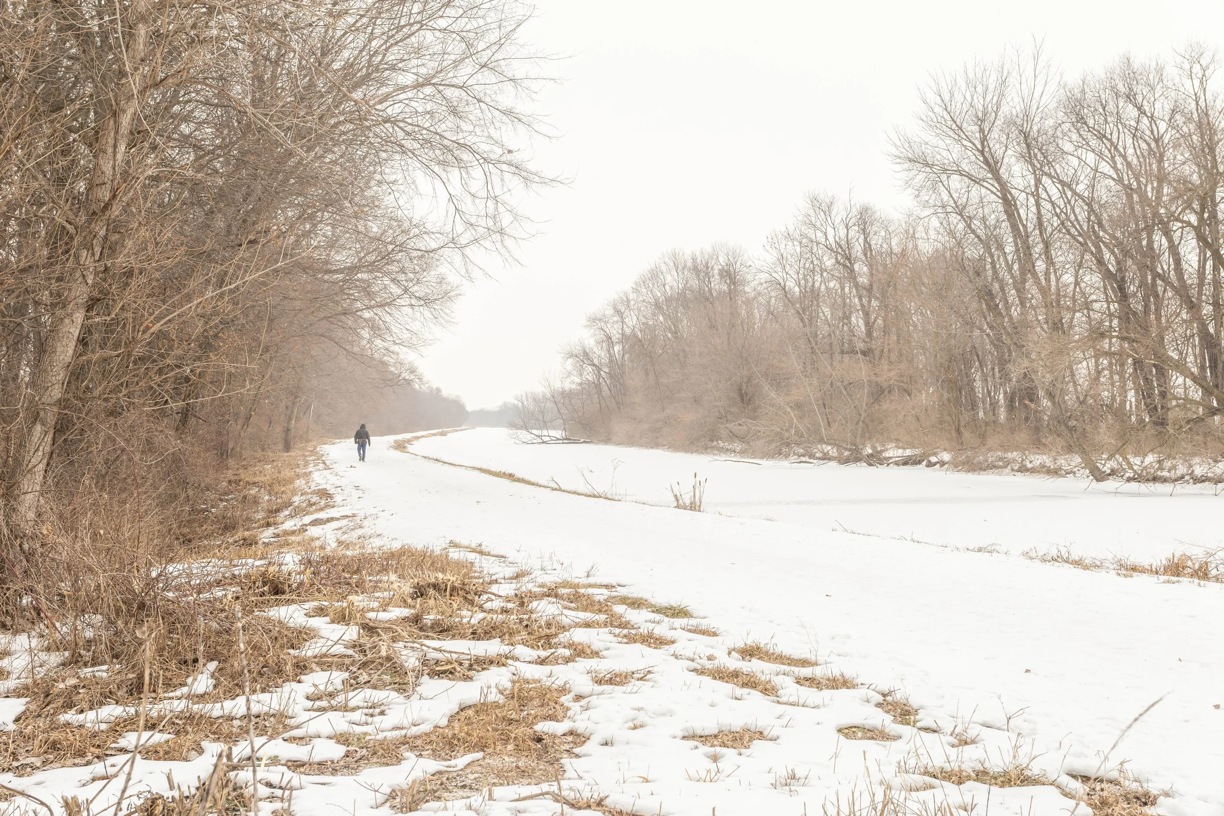 A person walking along a snow-covered trail beside a frozen river with leafless trees on either side on a cloudy winter day.