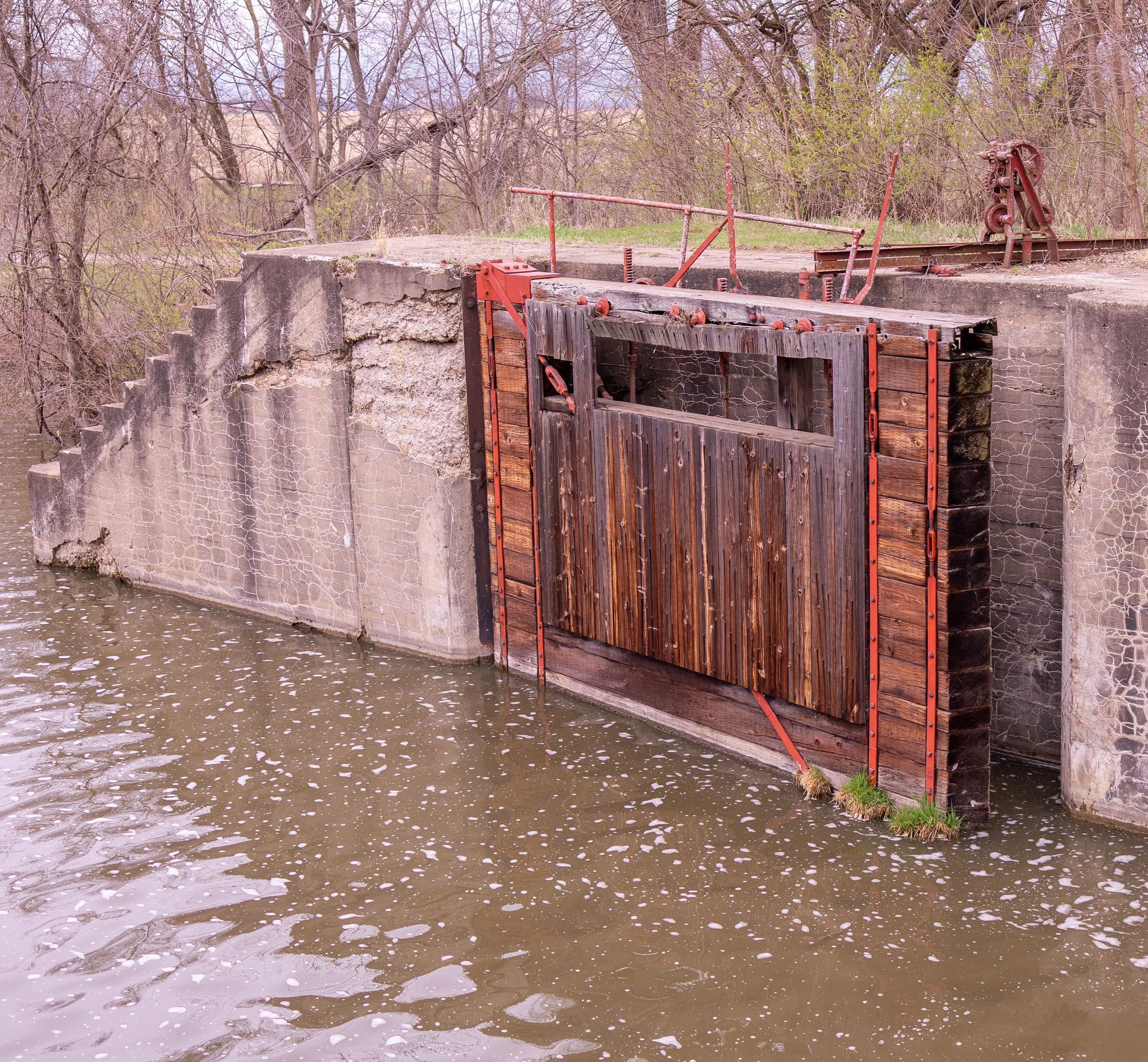 Old wooden water gate with a raised door in a concrete dam, surrounded by water and leafless trees.