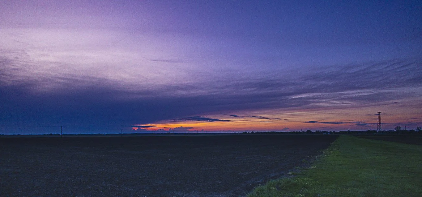 A vast open field during sunset with a colorful, cloudy sky and distant power lines.