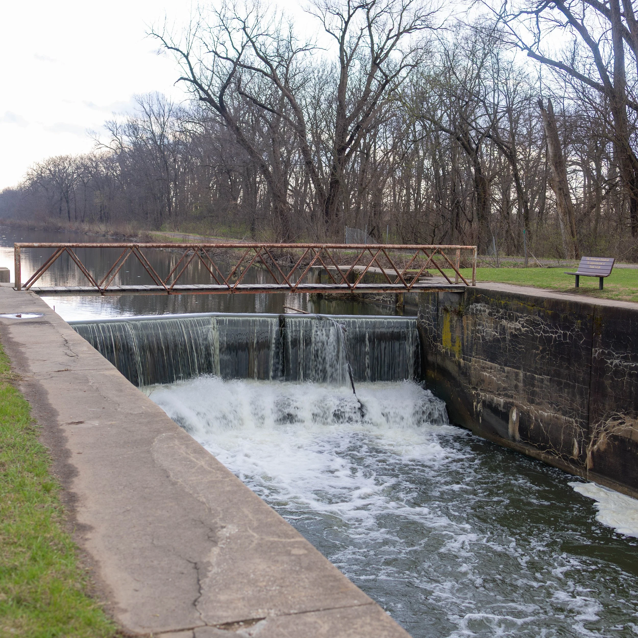 A small urban waterfall and dam with a rusted metal footbridge across it, surrounded by leafless trees and a park bench on a cloudy day.
