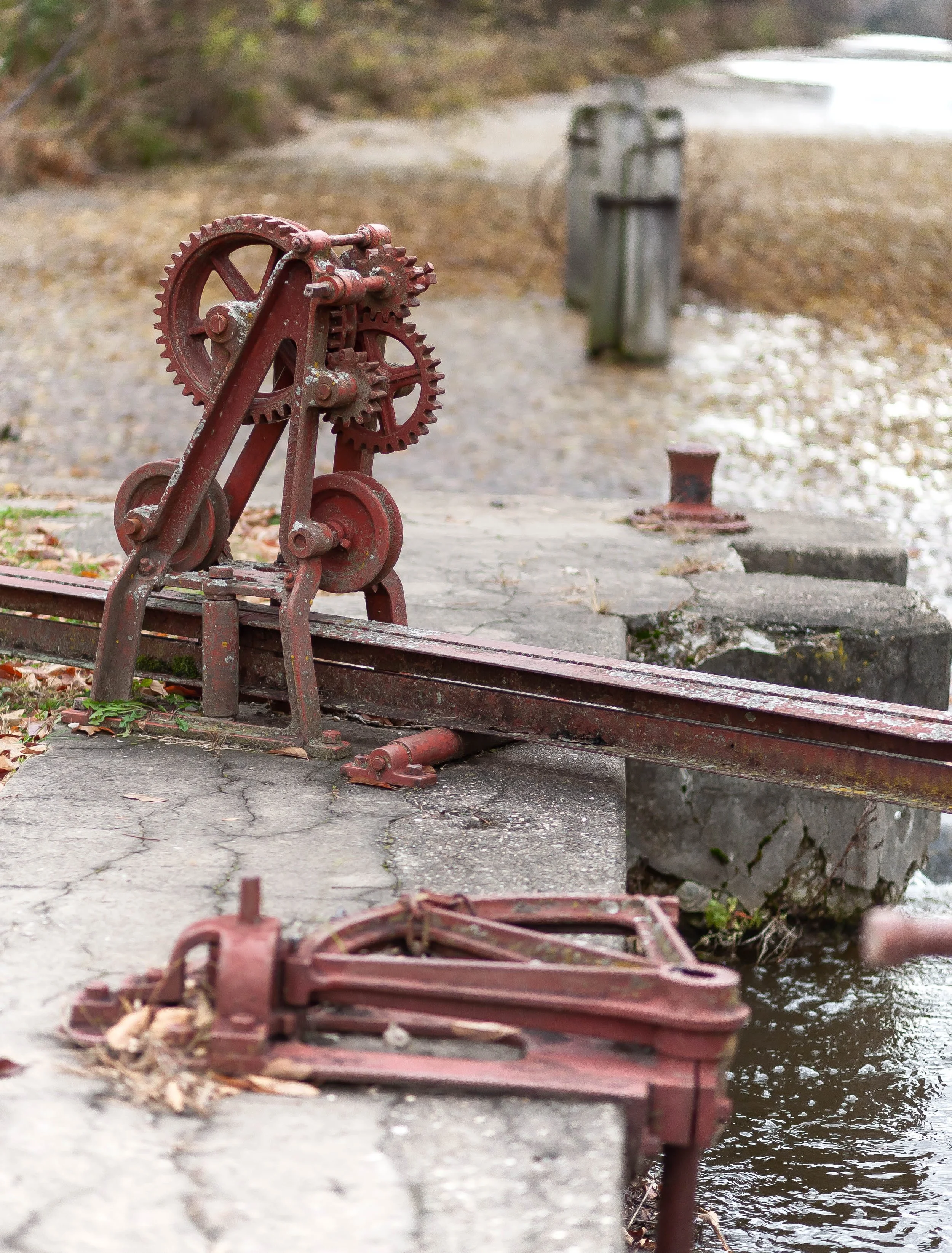 Old rusty mechanical equipment on a concrete dock beside a body of water, with background trees and pebbles.
