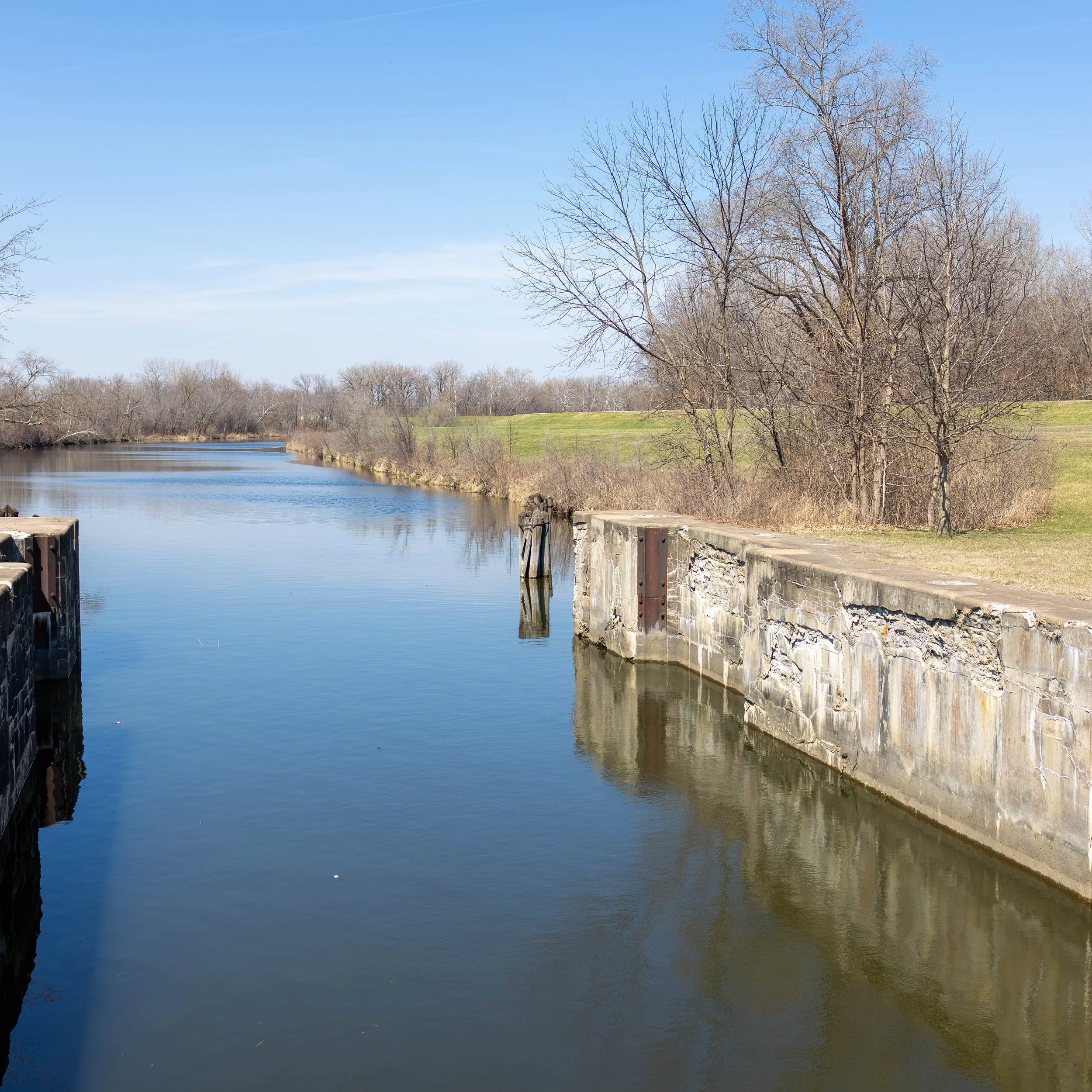 A calm river runs alongside an old stone and concrete dam with leafless trees and green grass in the background, under a clear blue sky.