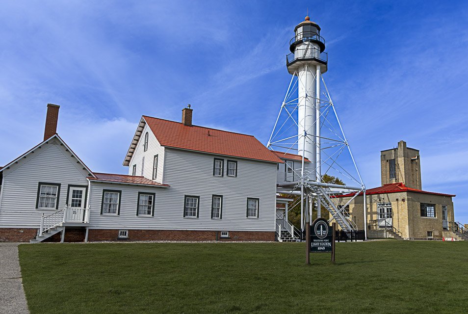 A white lighthouse with a black top and a red roof surrounded by two buildings, one with a red roof and the other with a beige stone exterior, on a grassy area under a blue sky.