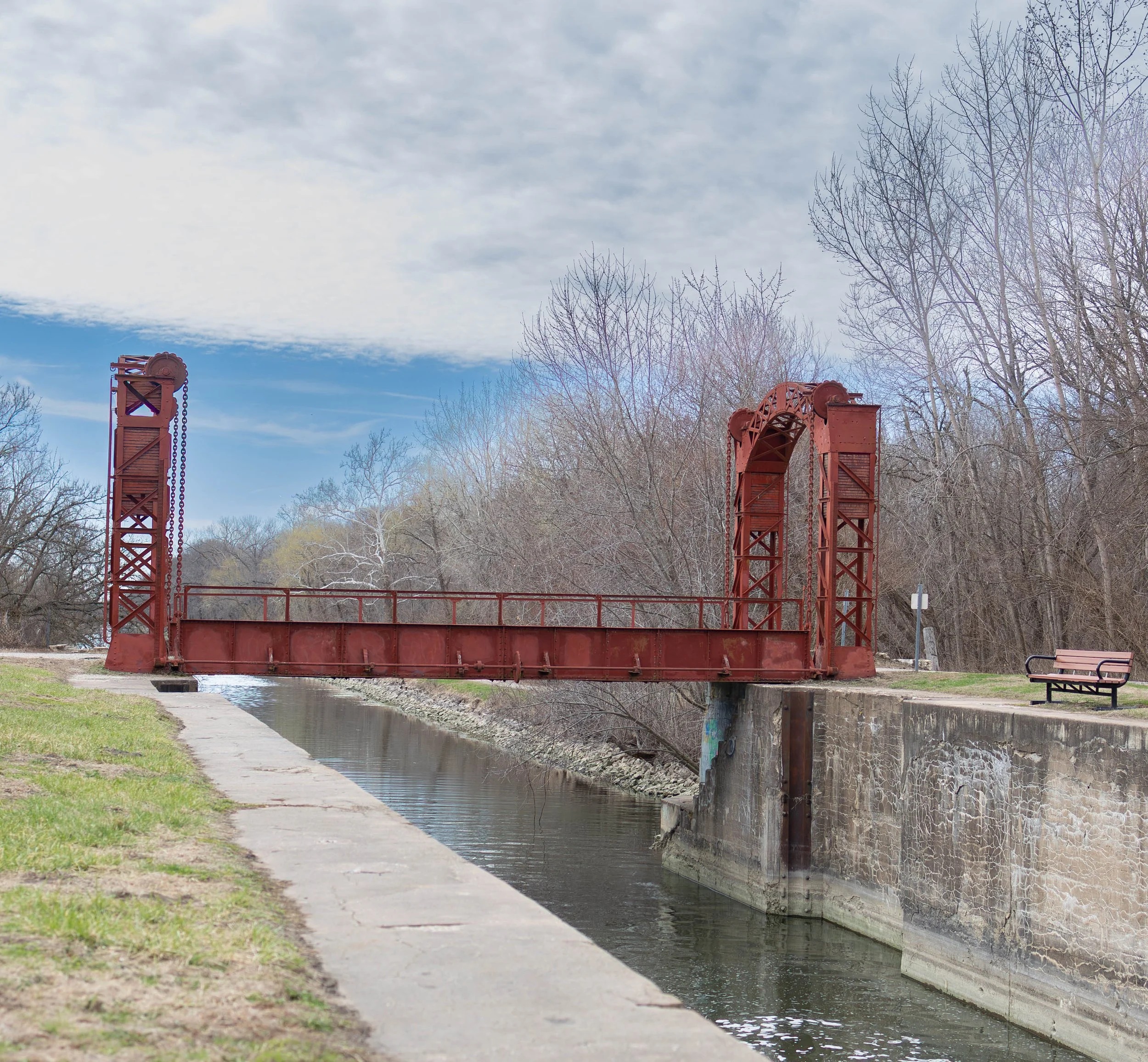 Old red metal bridge over a narrow canal with leafless trees in the background, a grassy area, a park bench, and a partly cloudy sky.