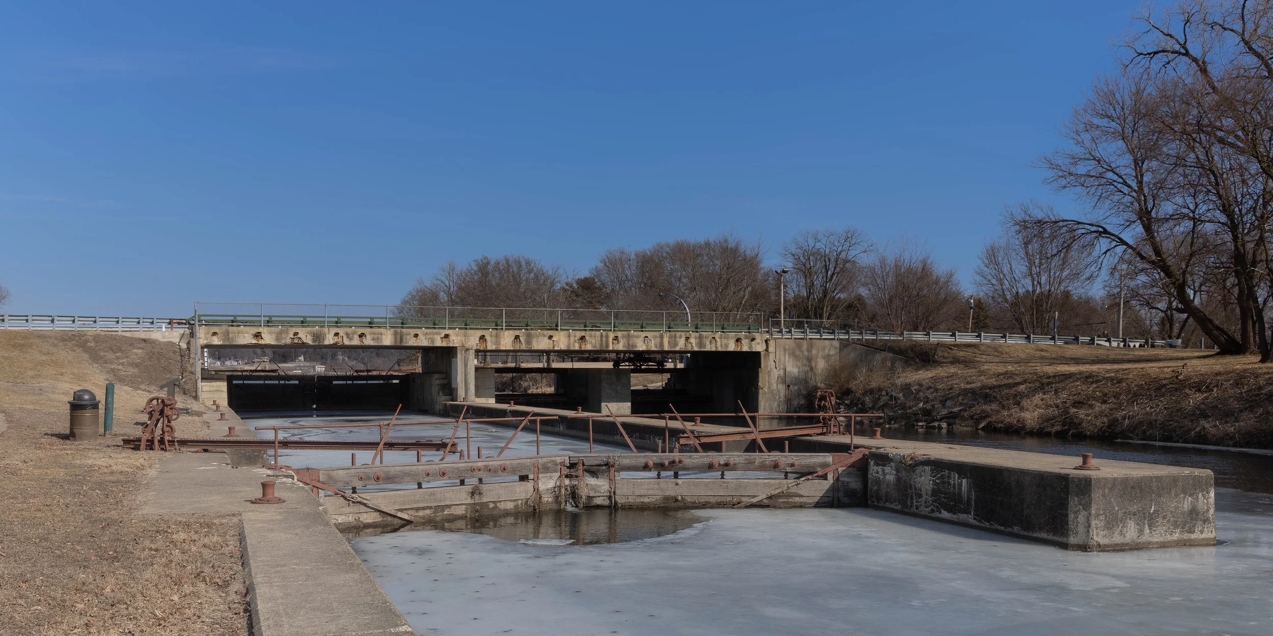 A dam structure on a river with a concrete wall and metal railings, closed off and submerged in ice and water, under a clear blue sky with leafless trees in the background.