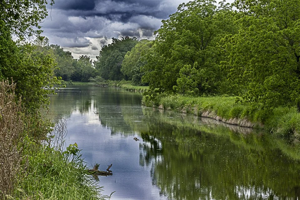 A calm river flowing through a lush green landscape with trees on both sides and a cloudy sky overhead.