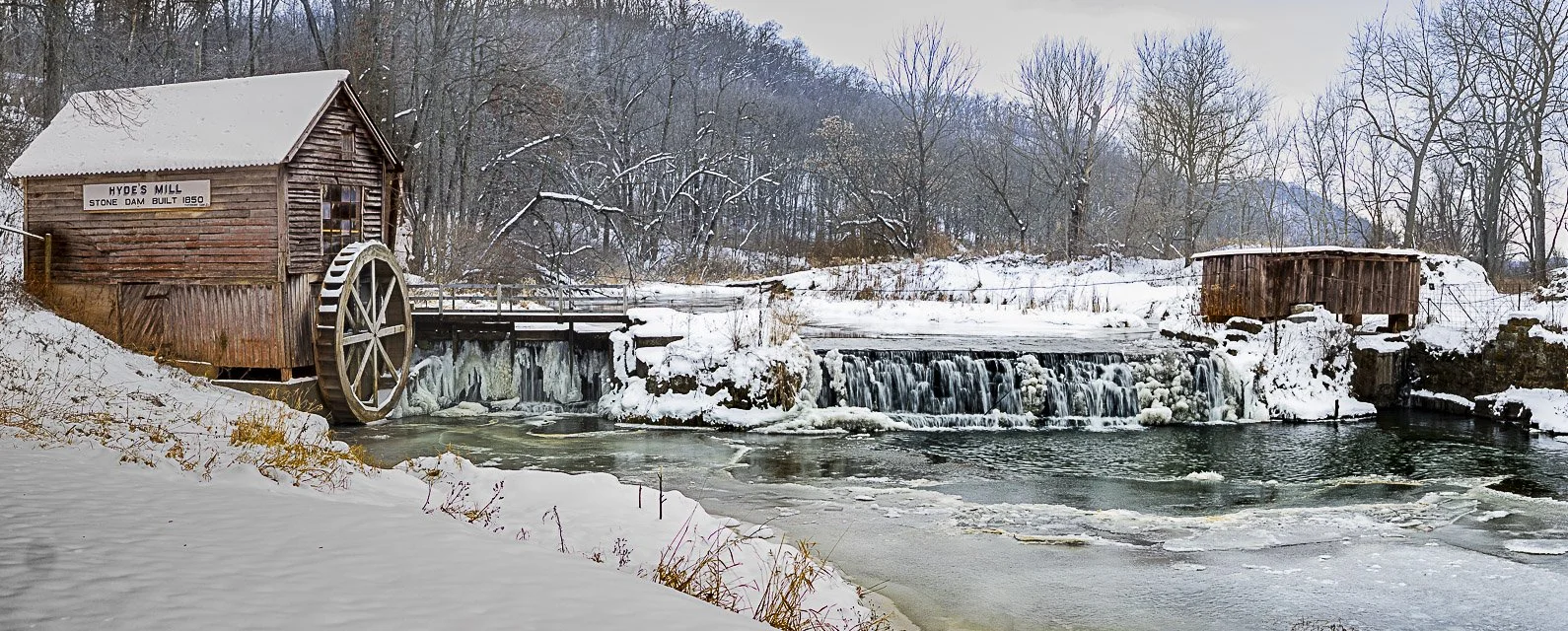 Snow-covered wooden watermill with a paddle wheel on the edge of a frozen river, surrounded by leafless trees in a winter landscape.
